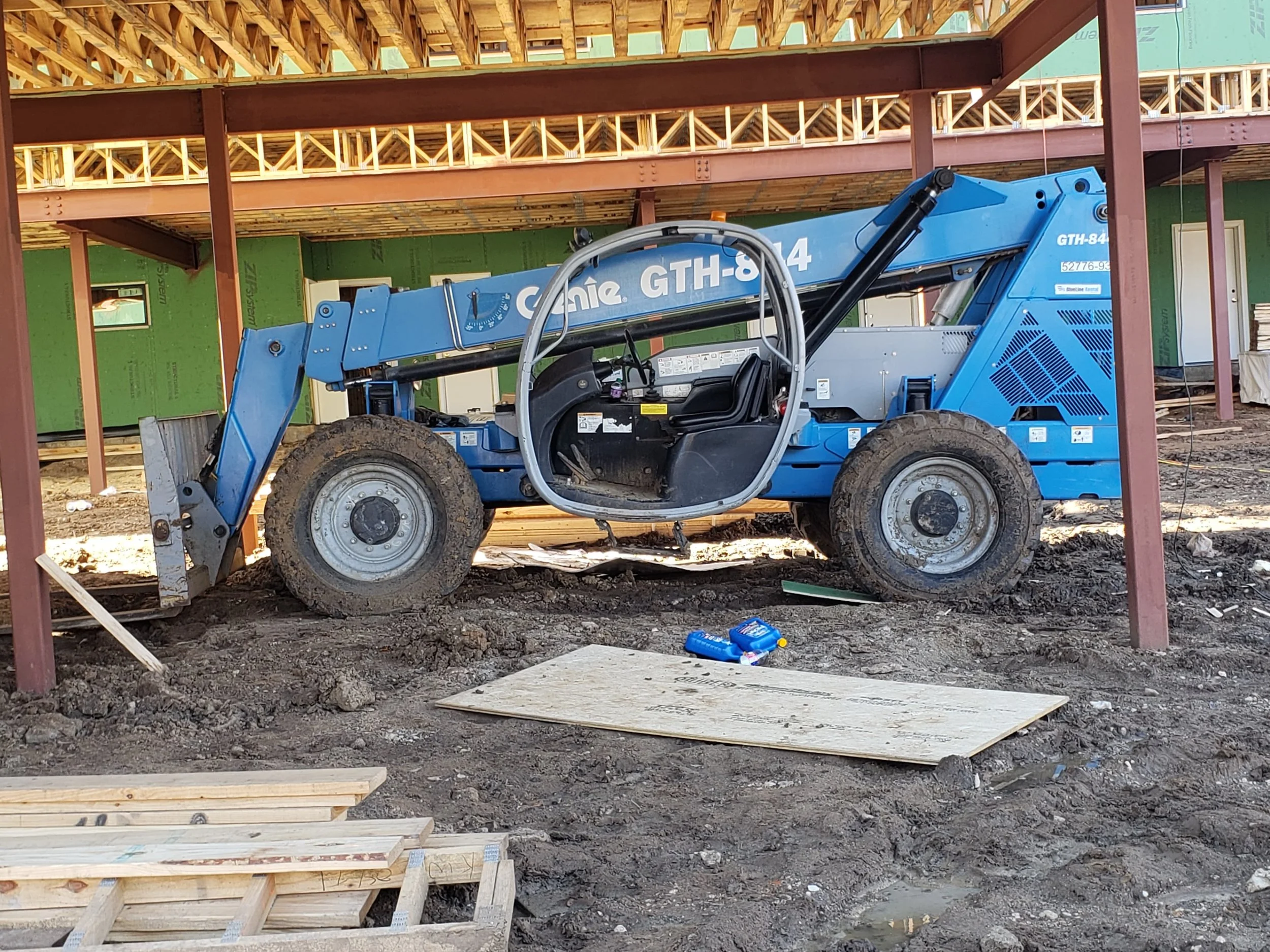 Construction site with a blue Genie GTH-844 telehandler forklift at work, with wooden framing and unfinished building structure in the background.