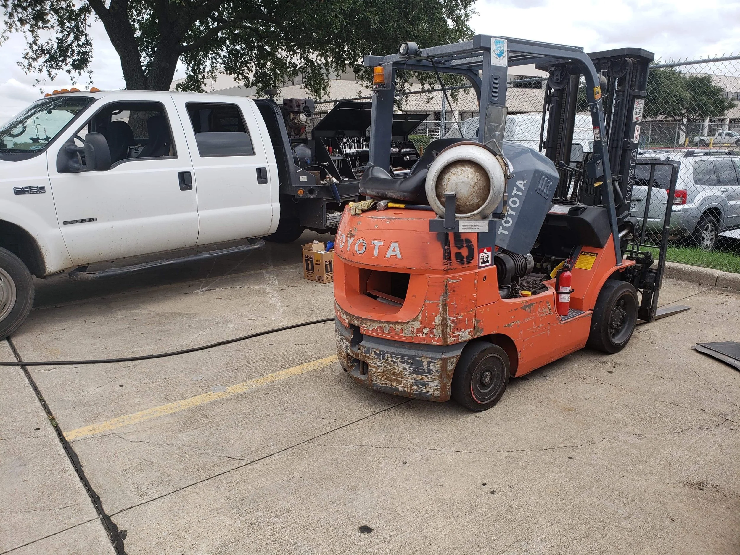 A white pickup truck is being serviced, with its bed open and a toolbox inside, parked next to a Toyota forklift with rust and dirt, in a parking lot with other vehicles and a chain-link fence.