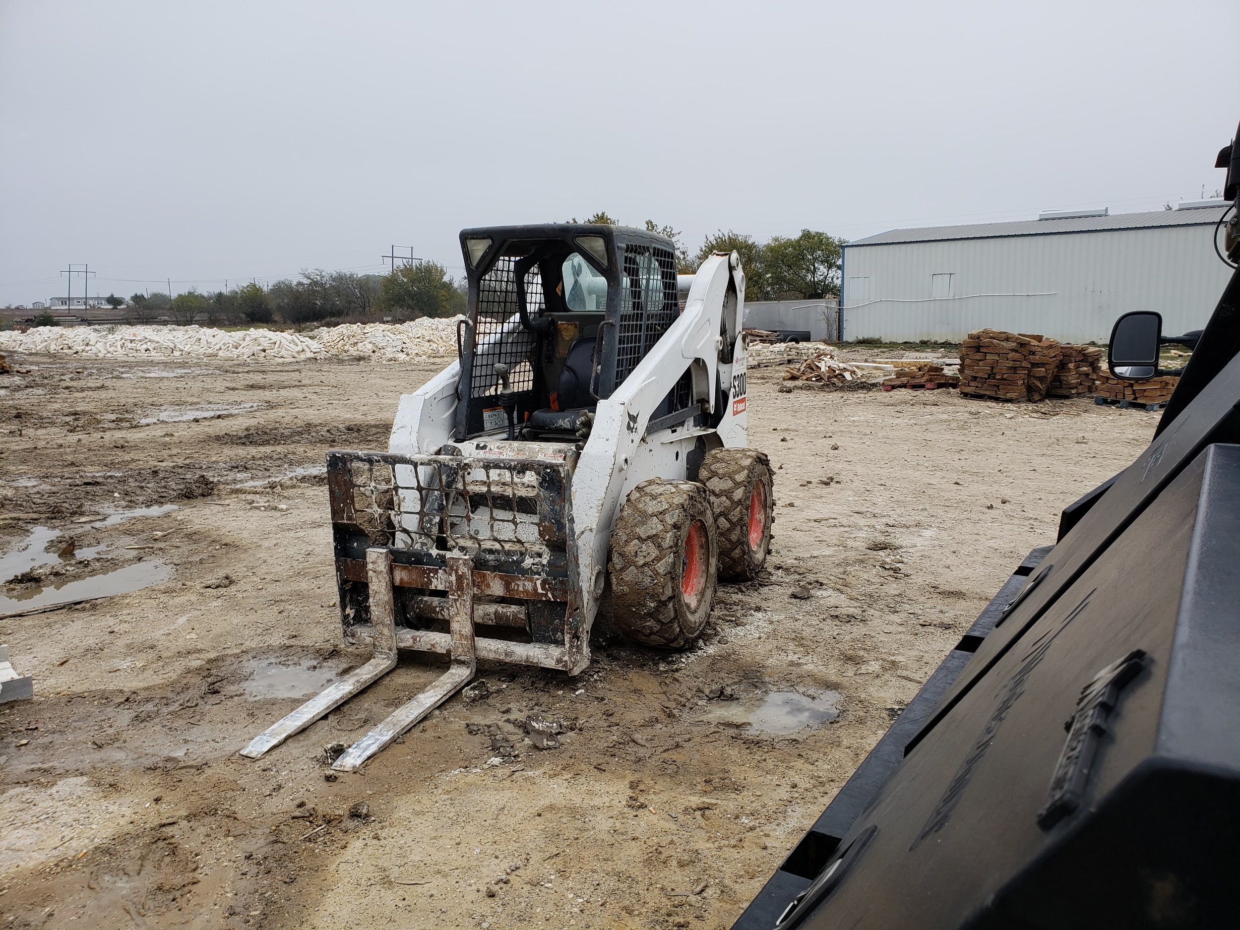 A construction site with a skid-steer loader, dirt ground, stacked bricks, and a large white building in the background under an overcast sky.