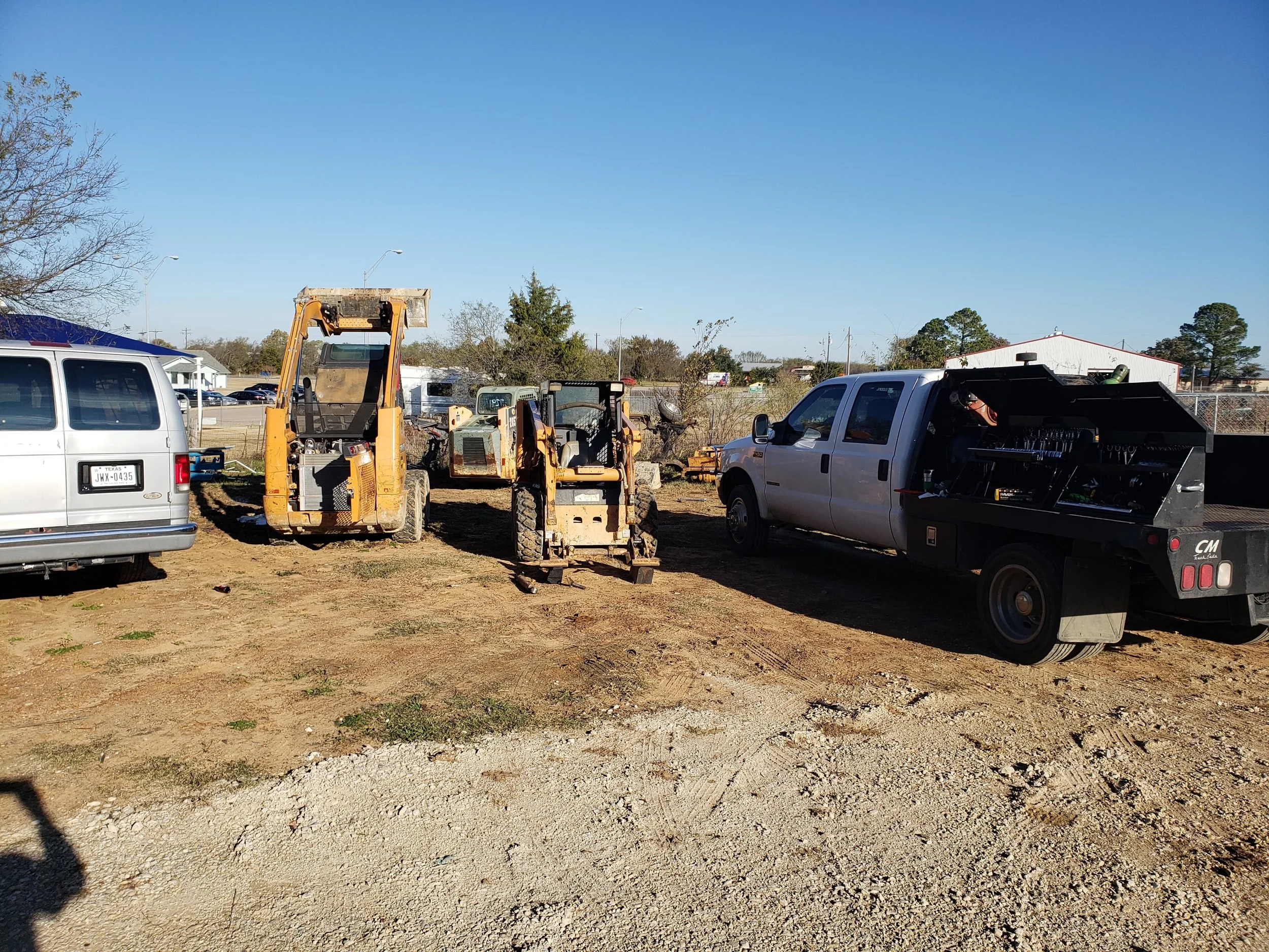 Construction site with a yellow skid-steer loader, a white van, and a service truck parked on dirt ground under a clear blue sky. There are trees and buildings in the background.