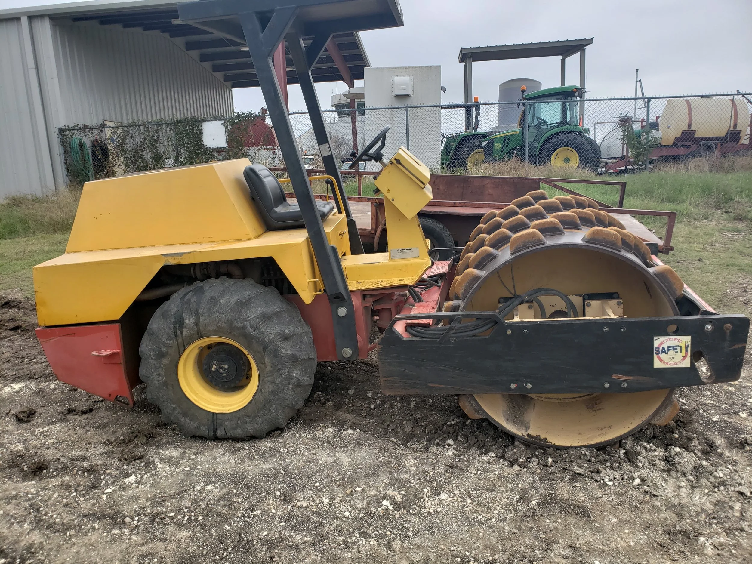 A yellow road roller with black rubber tires parked on dirt ground, with a wooden trailer attached, in an outdoor area with a chain-link fence and farm equipment in the background.
