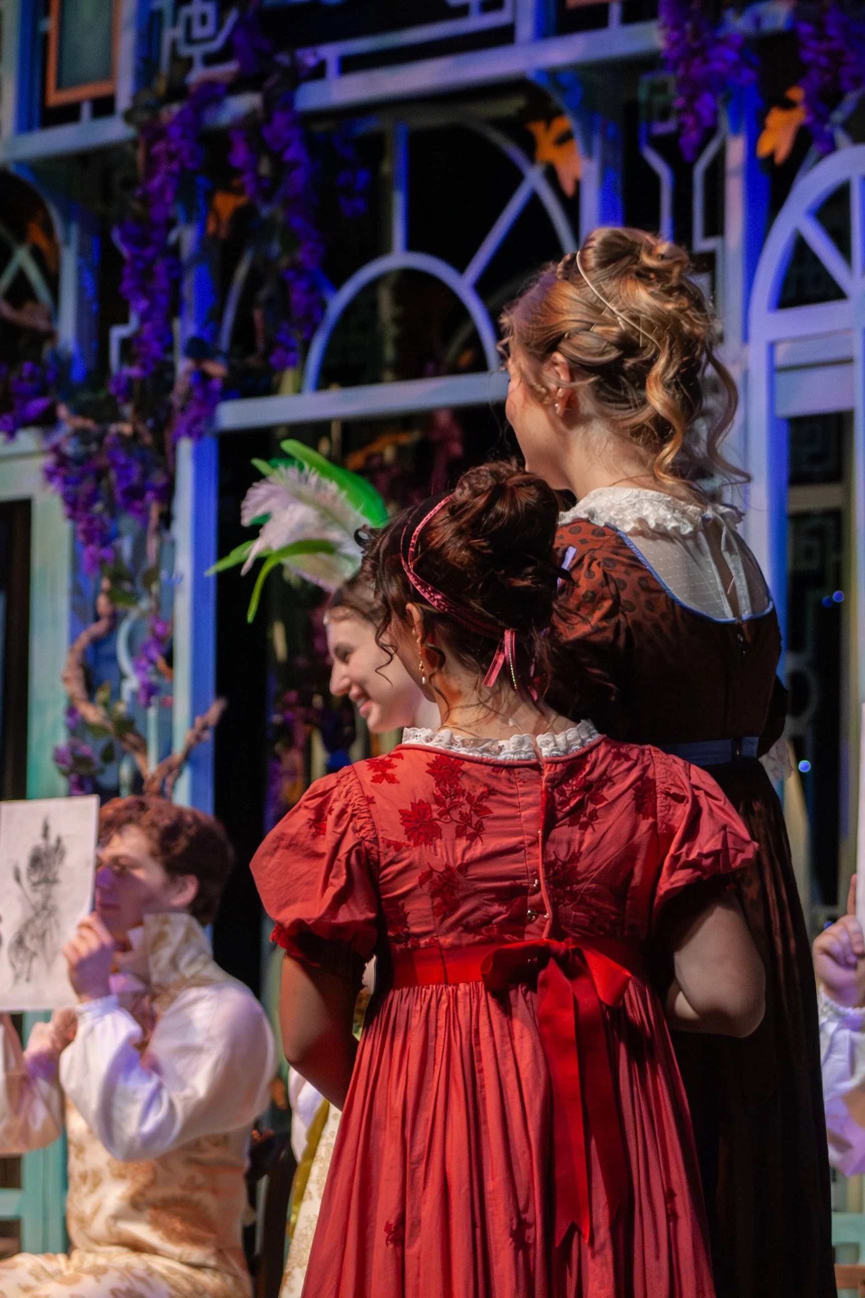Group of people in historical costumes at a performance or event, with colorful stage decorations and flowers in the background.