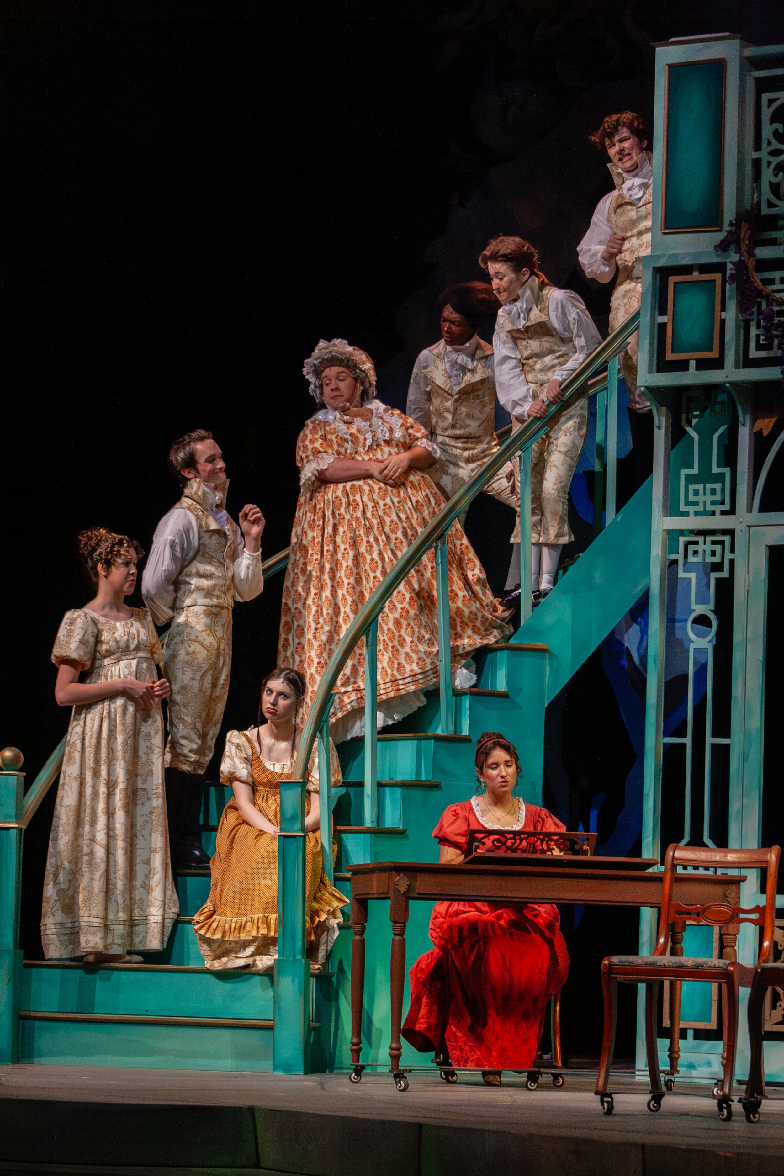 Actors in period costume performing on stage, with a teal staircase and a woman playing a piano.