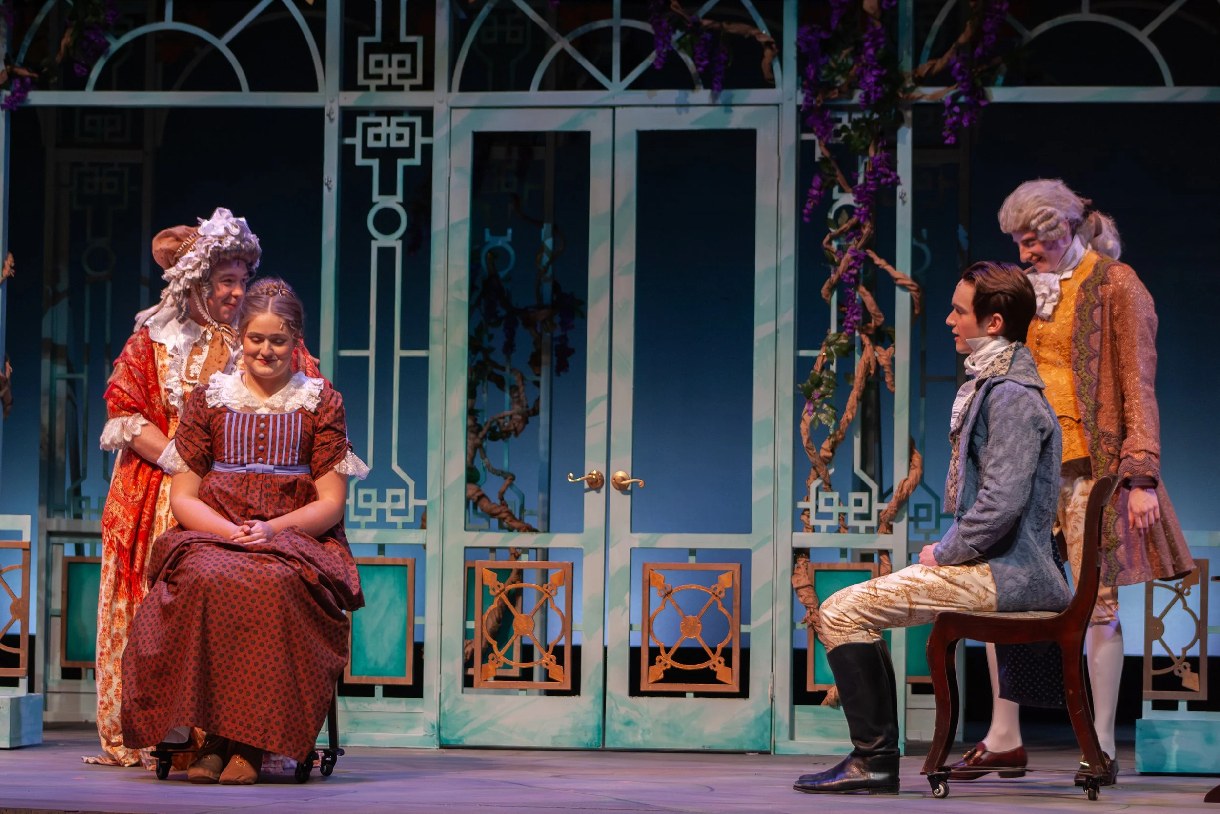 Actors performing on stage in period costumes, with a woman seated in a wheelchair. Behind them is a decorative set resembling a garden with purple flowers and blue-green fencing.