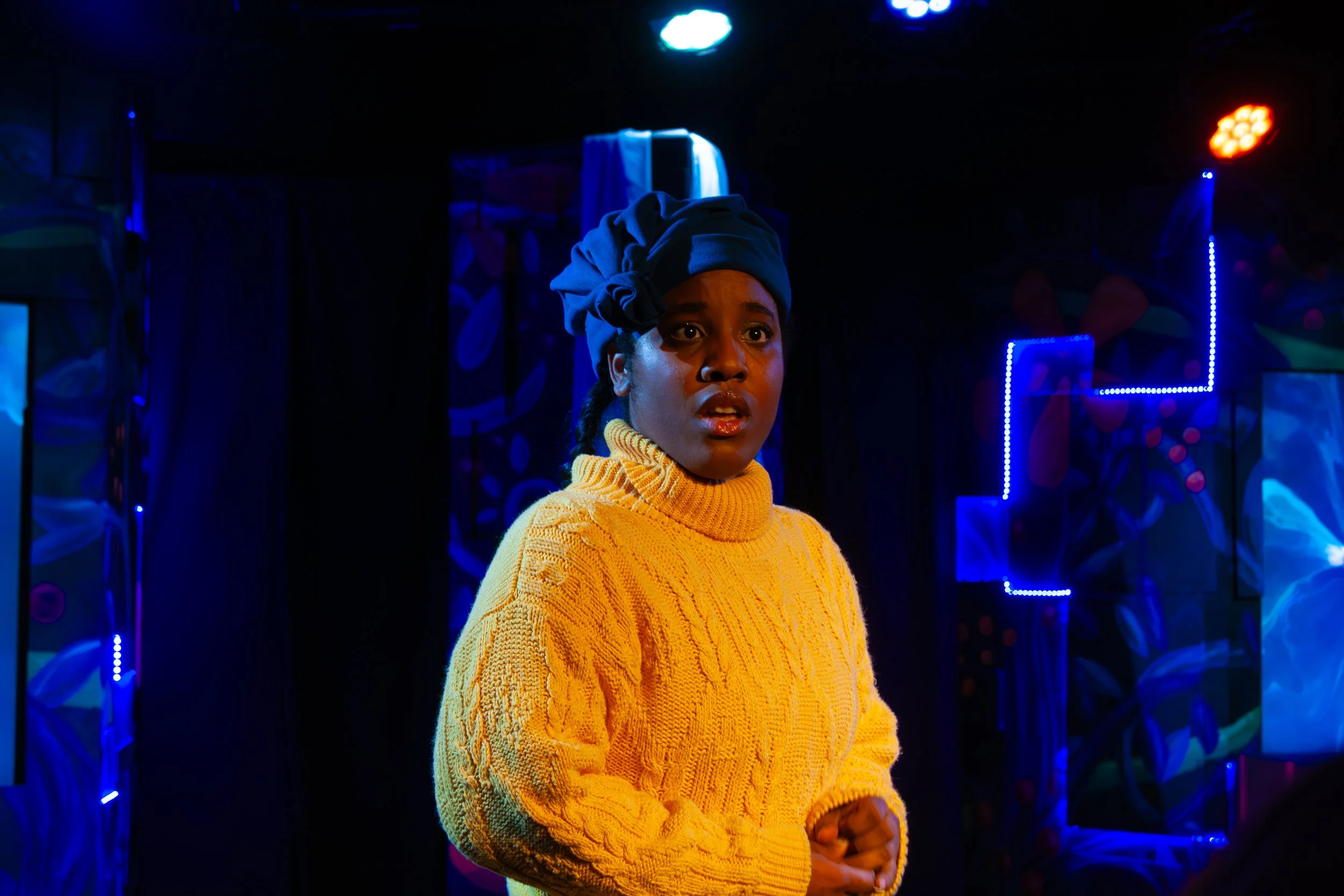 A woman with dark skin wearing a yellow knit sweater and blue headwrap, standing in a dark room with colorful neon lights and abstract wall designs.