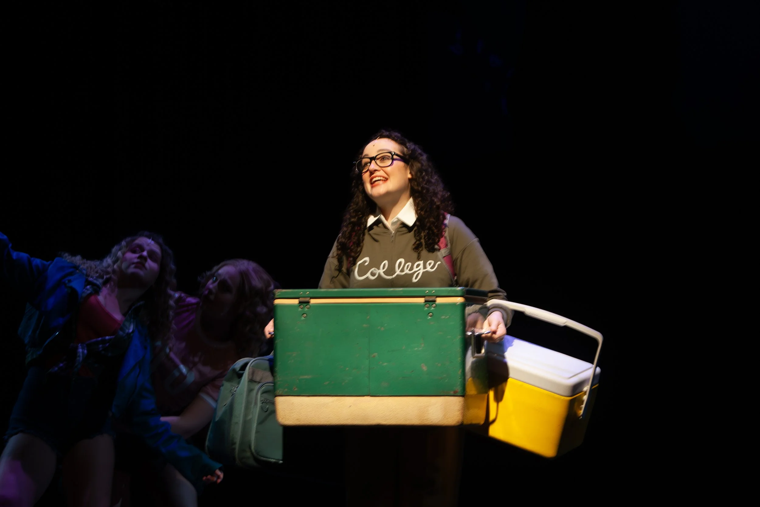 Young woman with glasses smiling, wearing a college hoodie, surrounded by others on a stage with dark background, pushing a cart with colorful storage containers.