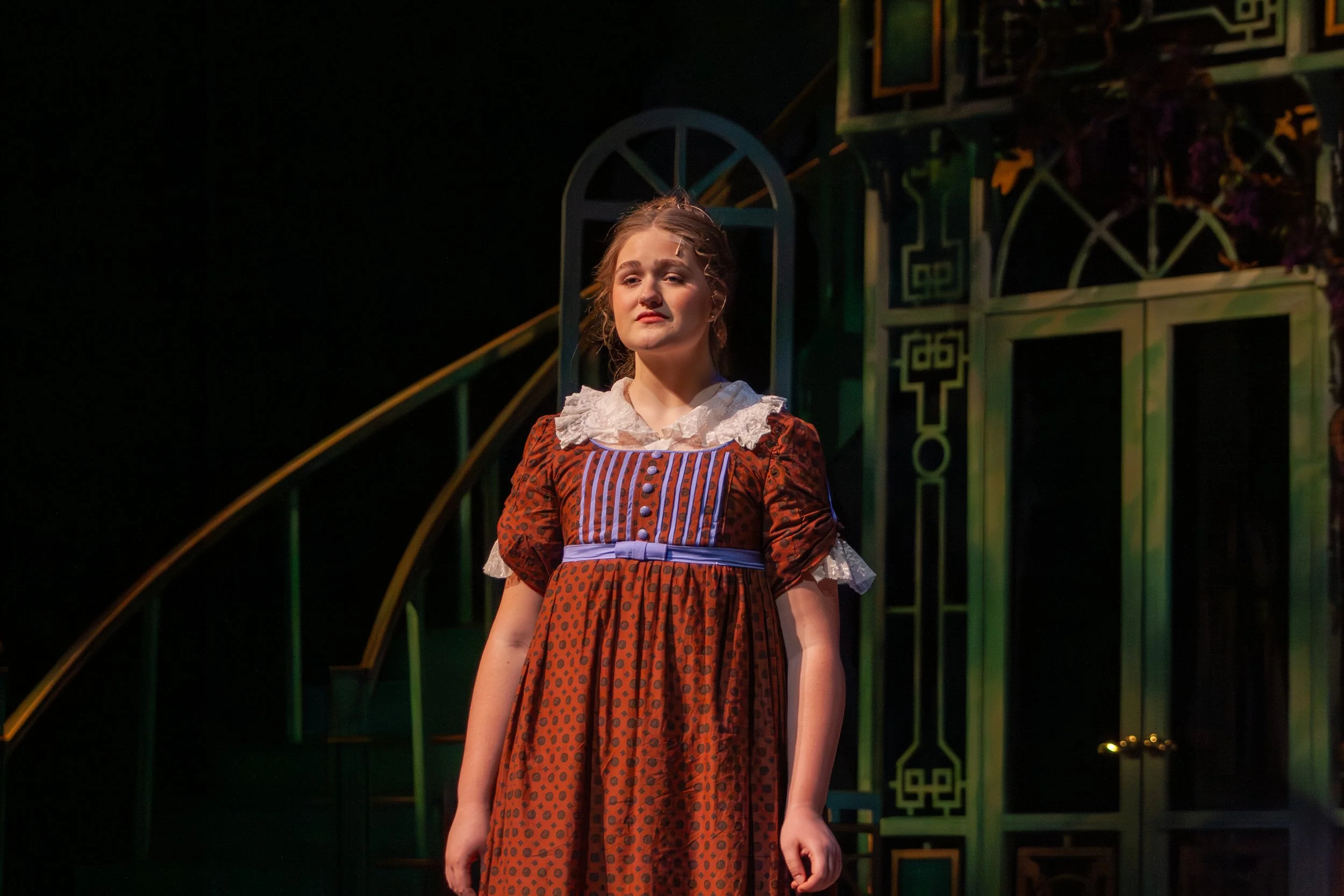 A young woman on stage wearing a vintage-style dress with a gathered waist and lace collar, standing in front of a decorative set with gothic arch motifs, a railing, and dark lighting.