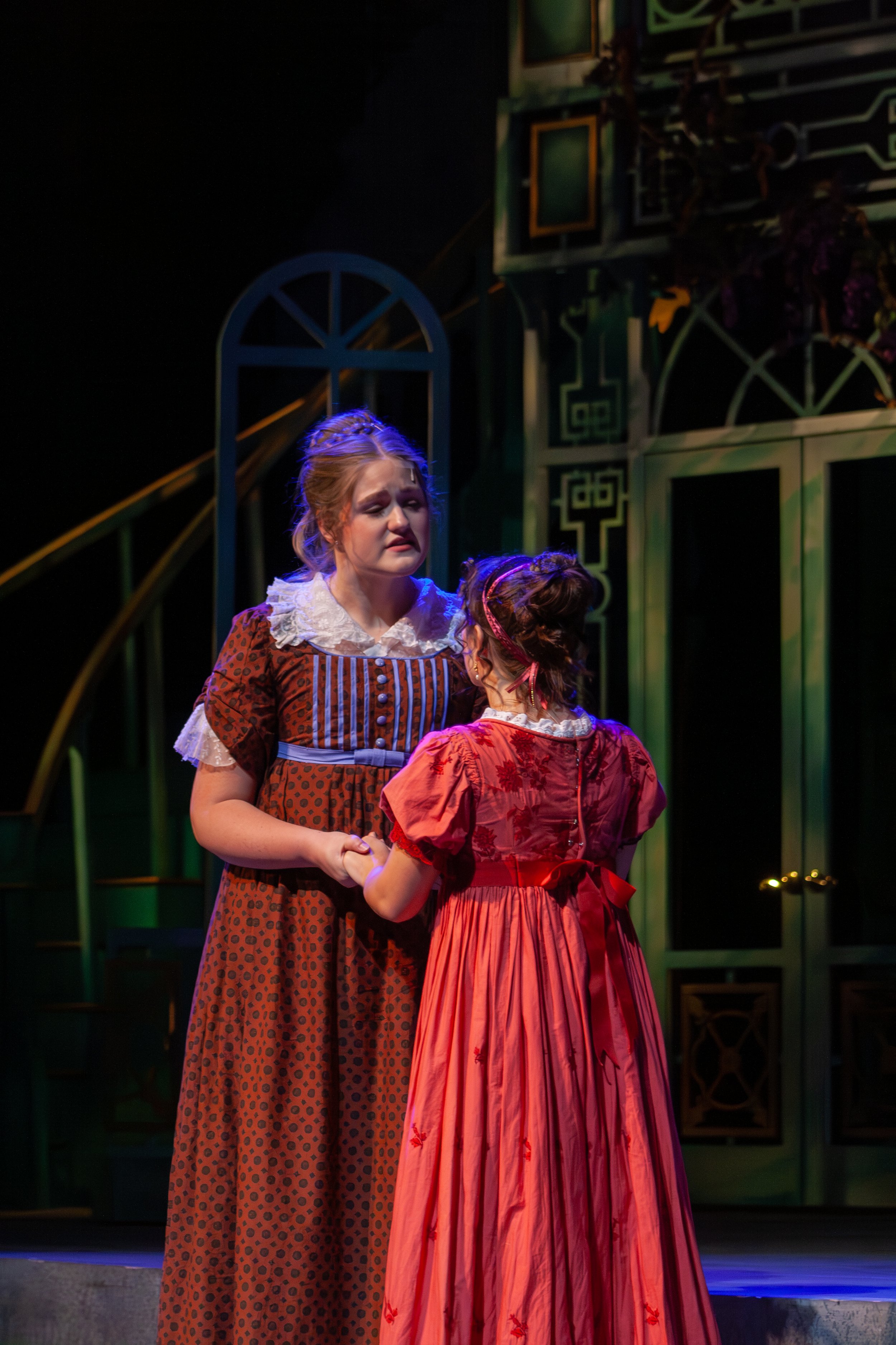 Two women dressed in period costumes are on stage, with one appearing distressed and the other comforting her. The background has theatrical set elements.