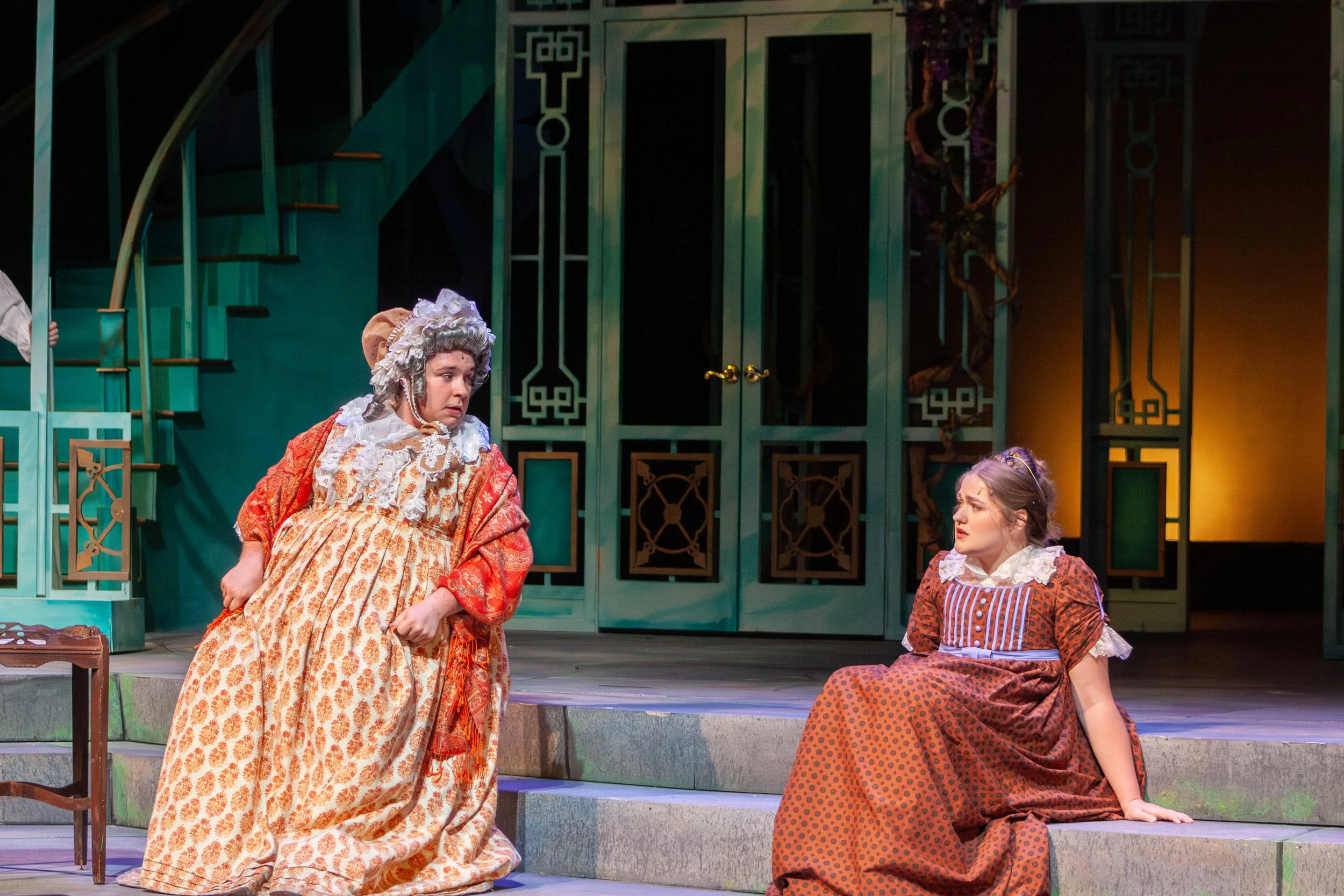 Three women in historical costumes performing on stage, with a teal and black set decor, including a staircase and decorative panels.