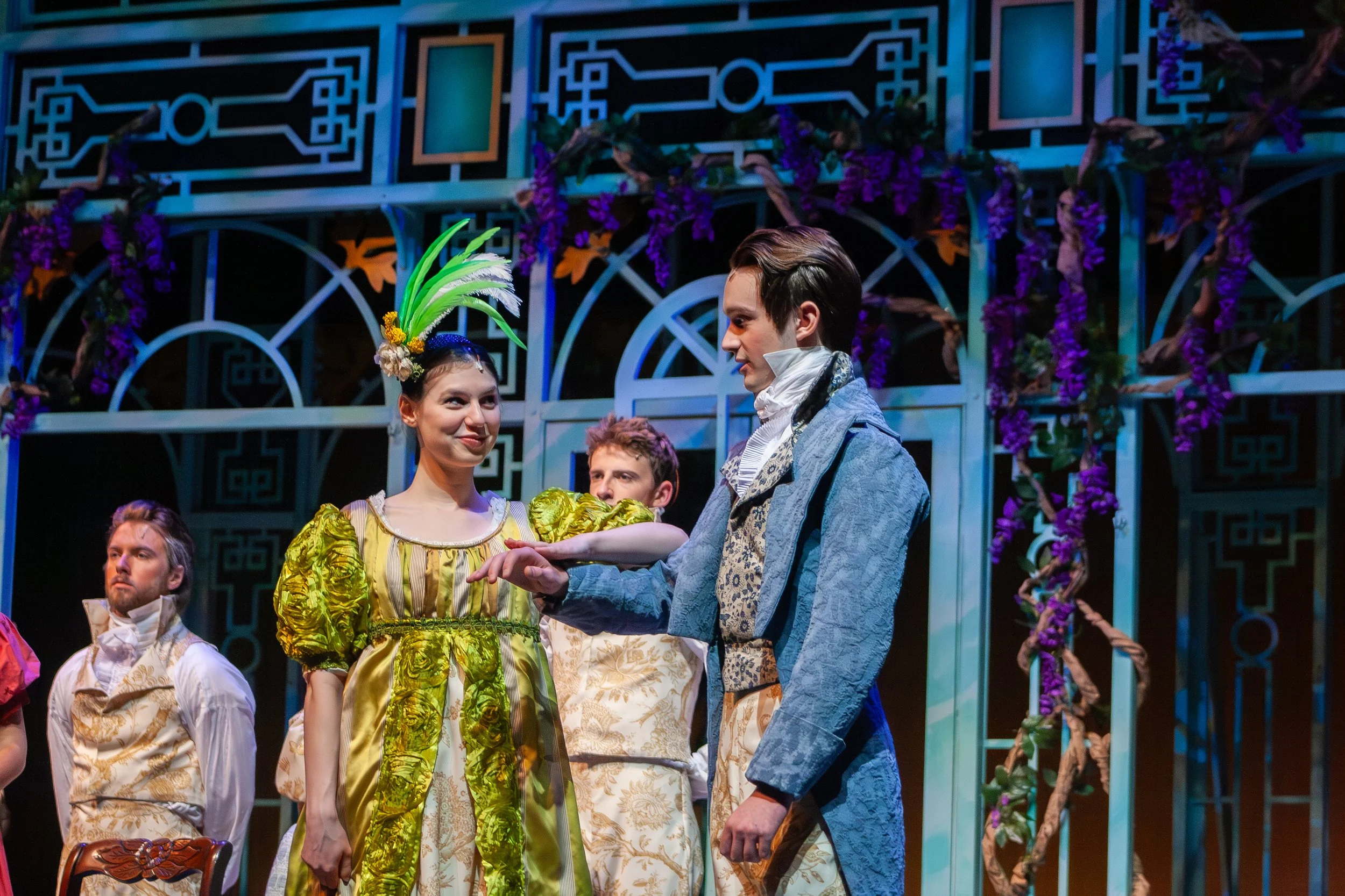Stage scene from a theatrical performance featuring actors in 18th-century costume. One woman in a yellow dress with green accents and a feathered headpiece stands in the center, smiling at a man in a blue coat. Others are seated or standing nearby, 