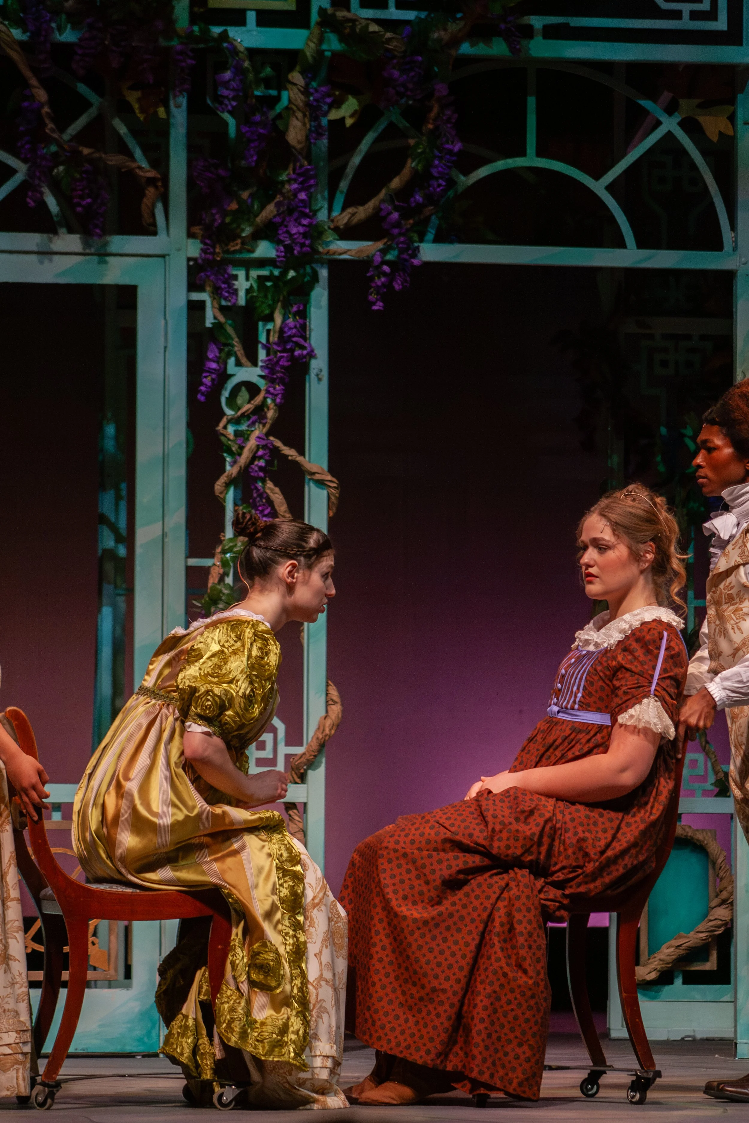Scene from a theatrical performance with actors dressed in historical costumes. Two women are seated, facing each other, with one wearing a yellow dress and the other an orange dress. Two men are standing nearby, dressed in period clothing. The backg