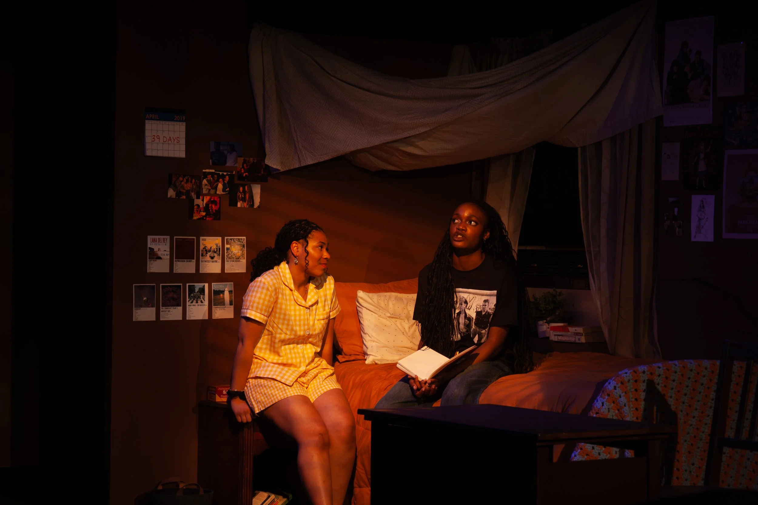 Two women sitting in a dark room, engaged in conversation. One woman is wearing a yellow checkered outfit, the other is in a black t-shirt. There are photos and posters on the wall behind them, and a calendar showing 39 days left in April 2019.