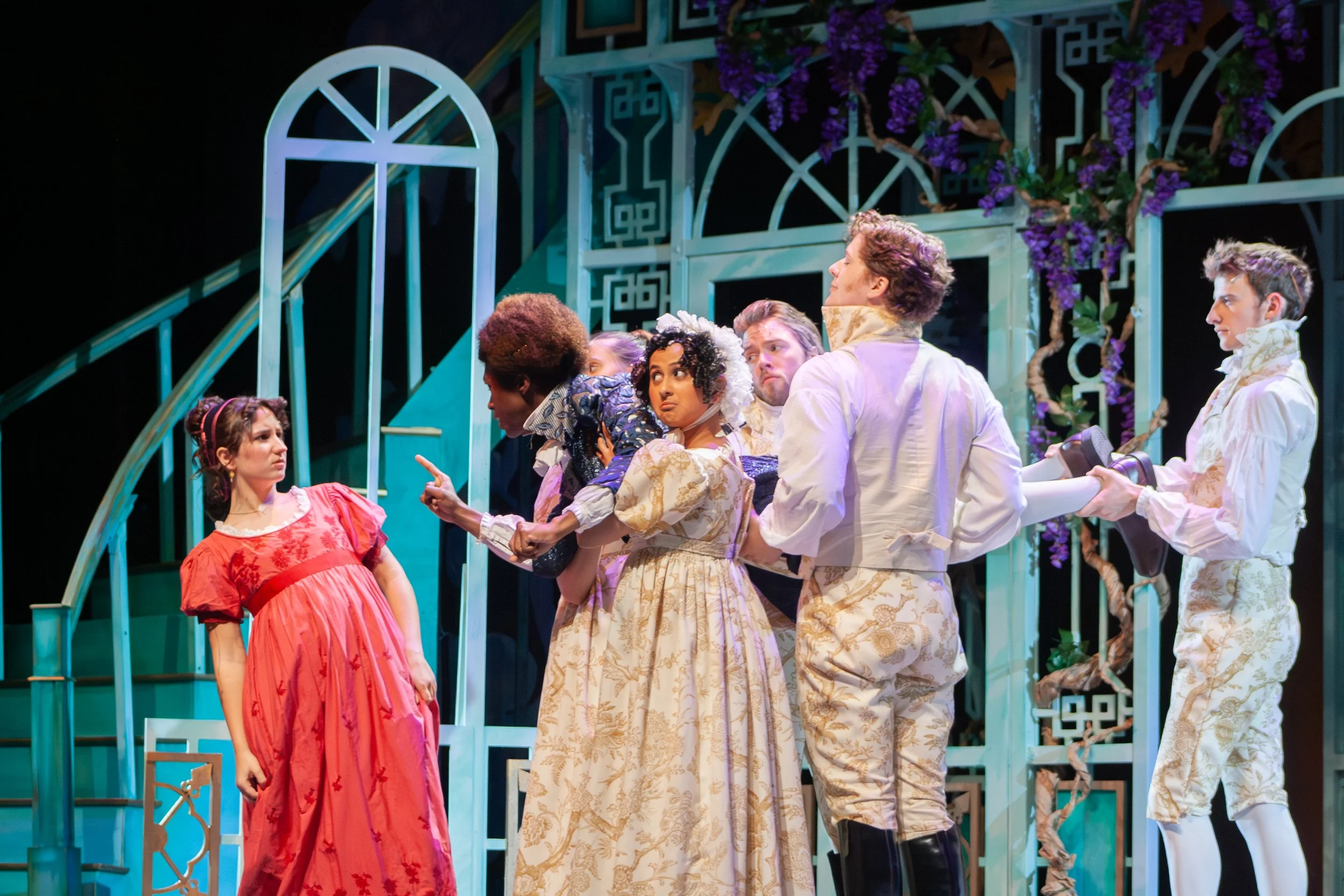 Actors on stage during a theatrical performance. The scene features a young woman in a red dress and several men in elaborate period costumes, with a decorative set background decorated with purple flowers.