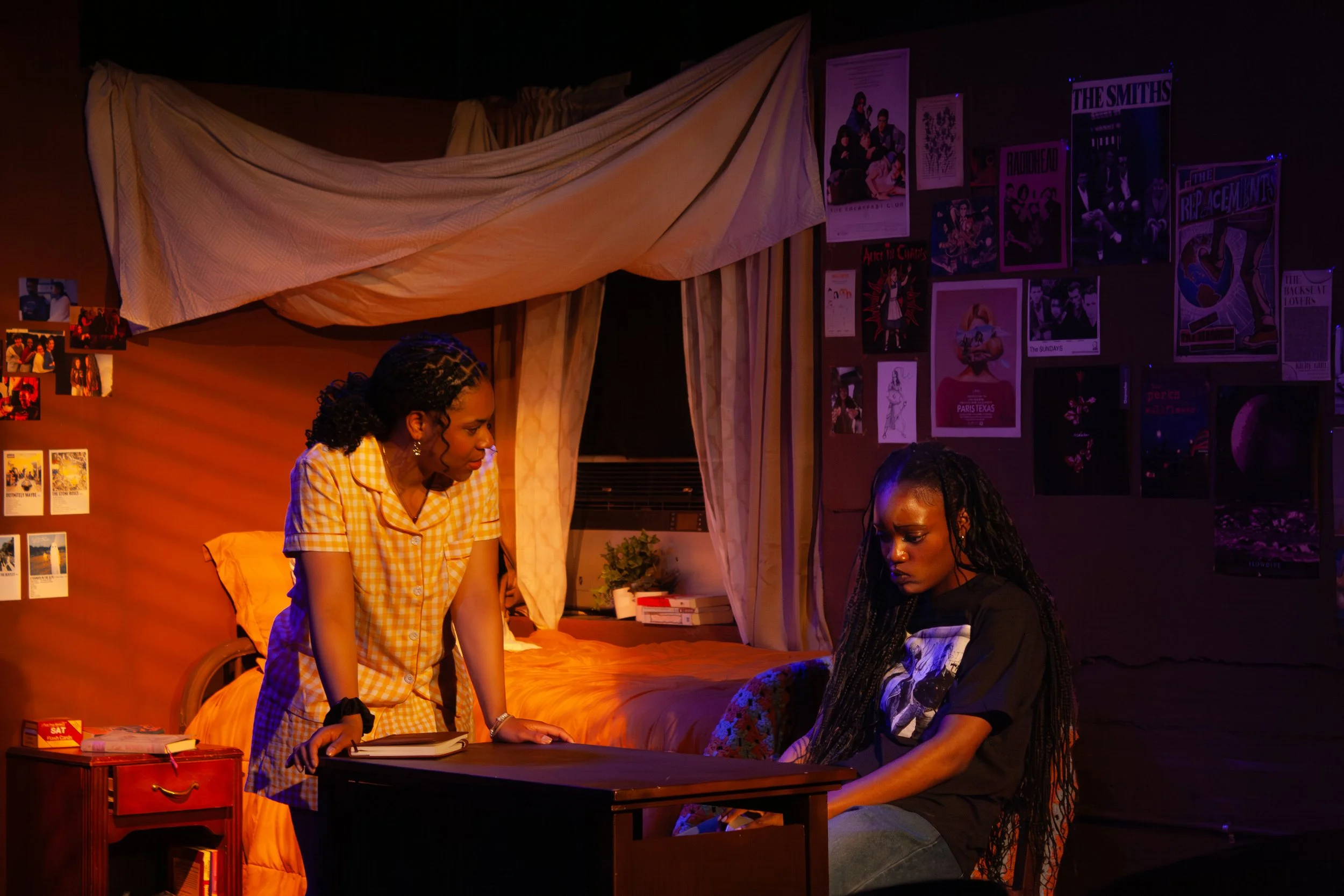 Two women in casual clothing and one woman with long braids sitting at a desk in a dimly lit room decorated with posters on the walls, a bed in the background, and a small red nightstand.