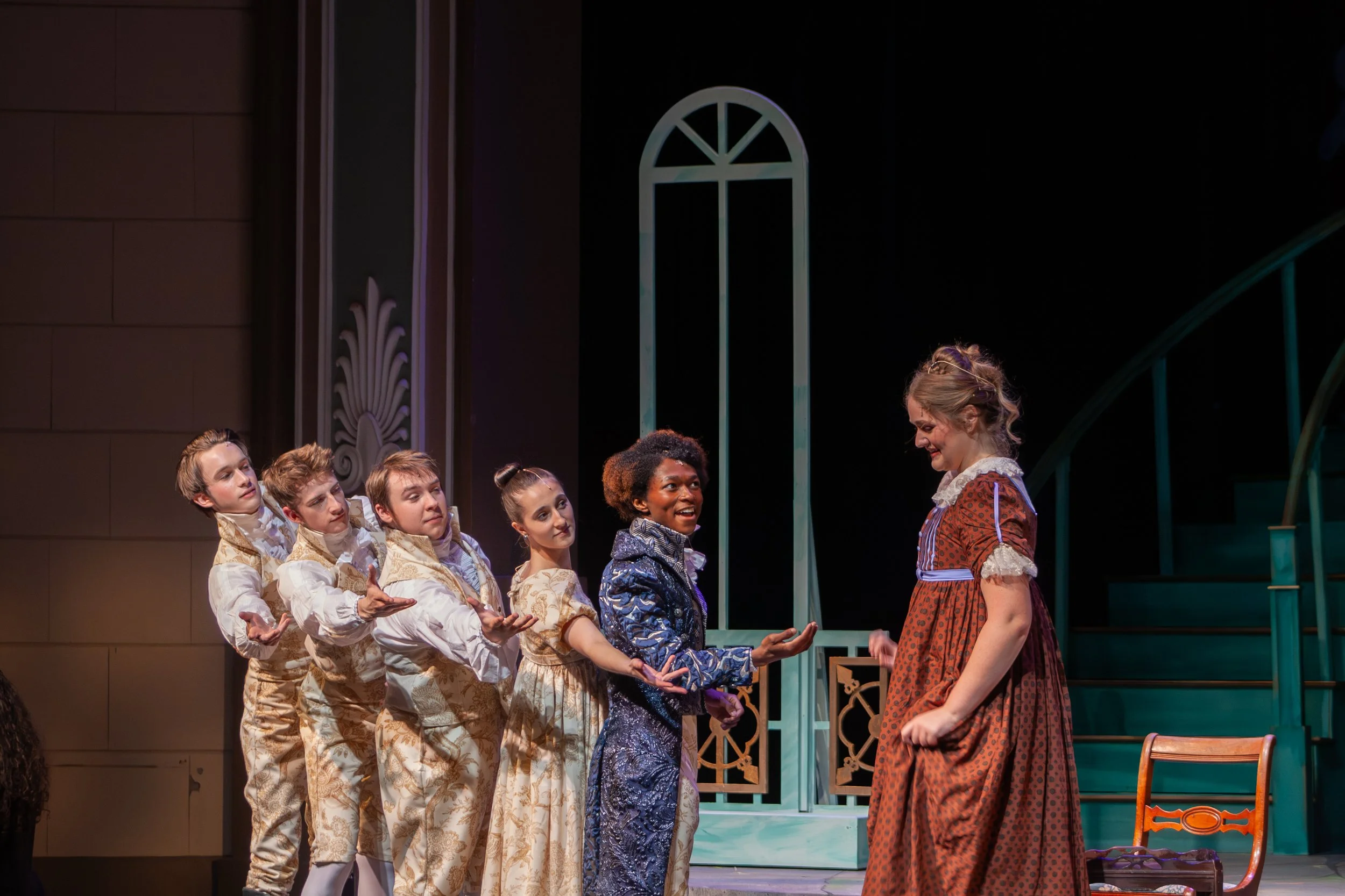 A group of children in elaborate costumes stand in a line on stage, interacting with a woman dressed in a period costume, with a backdrop resembling a house with arched windows and decorative elements.