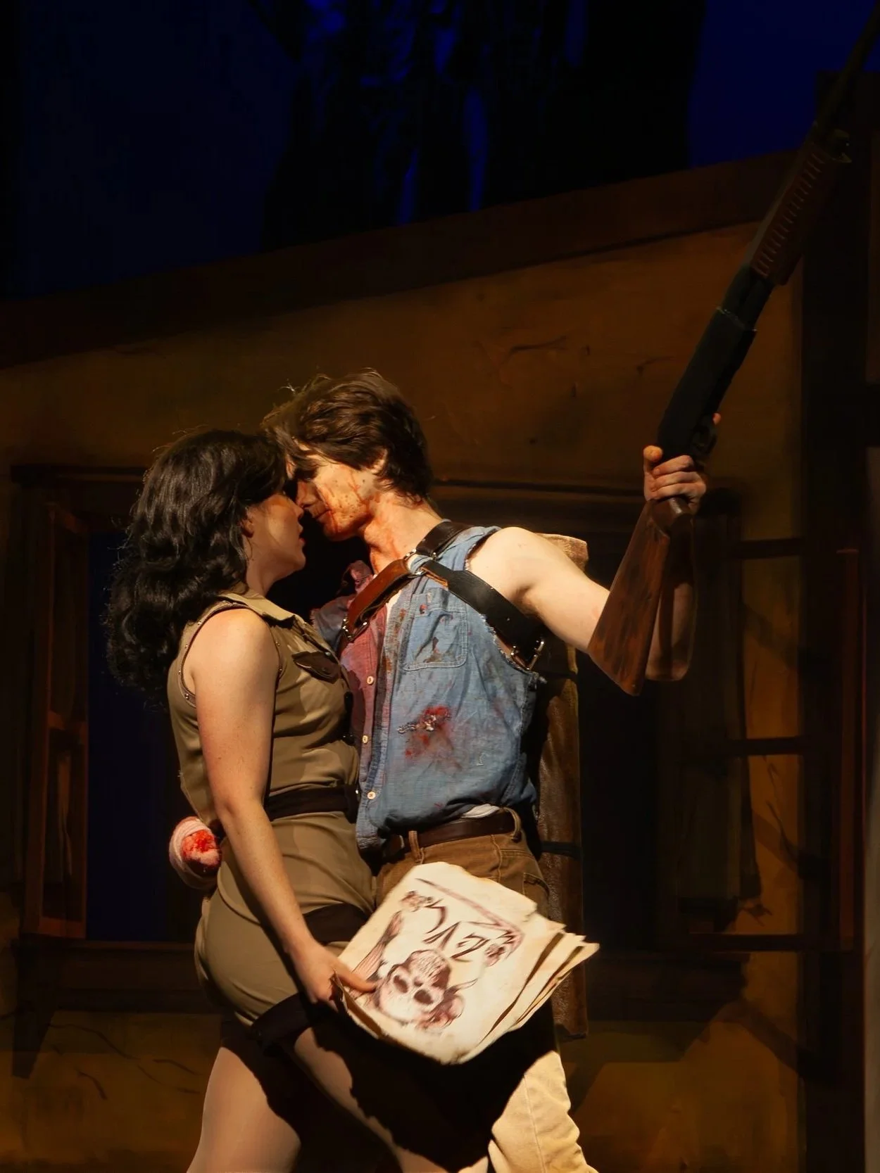 Two actors in a theatrical scene, one man with bloodstains and blood on his face holding a shotgun, and a woman in a tan uniform holding a newspaper with a dog illustration, standing closely with foreheads touching.