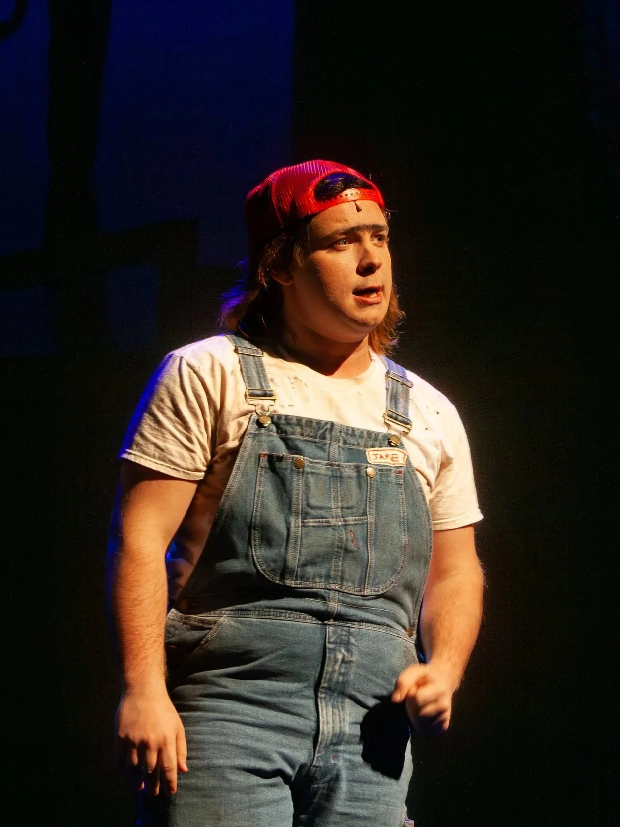 Young man with long hair wearing a red backward cap, white shirt with stains, and denim overalls, standing on stage with a serious expression, holding a microphone in his right hand, lit by stage lights.