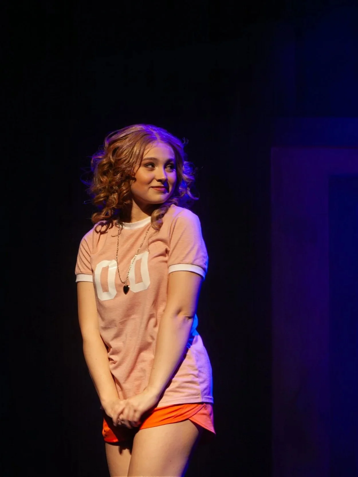 A young woman with curly blonde hair, wearing a pink t-shirt with the number 00 printed on it, and orange shorts, standing on a stage with purple and blue lighting, with a dark background.