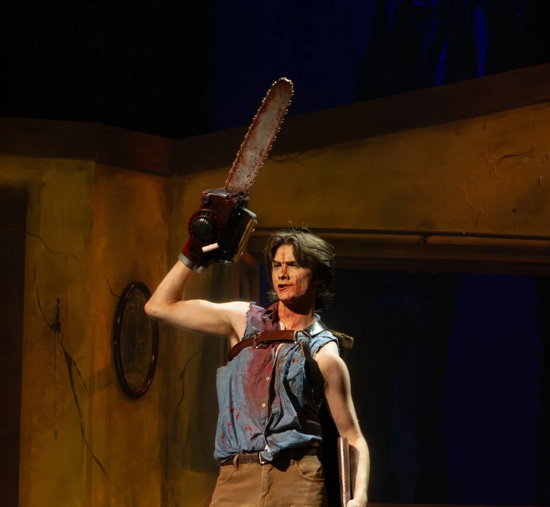 A young man with disheveled brown hair and a blood-stained face holding a large, bloodied chainsaw above his head. He is wearing a sleeveless shirt and beige pants, standing in a rustic room with wooden walls and a mirror.