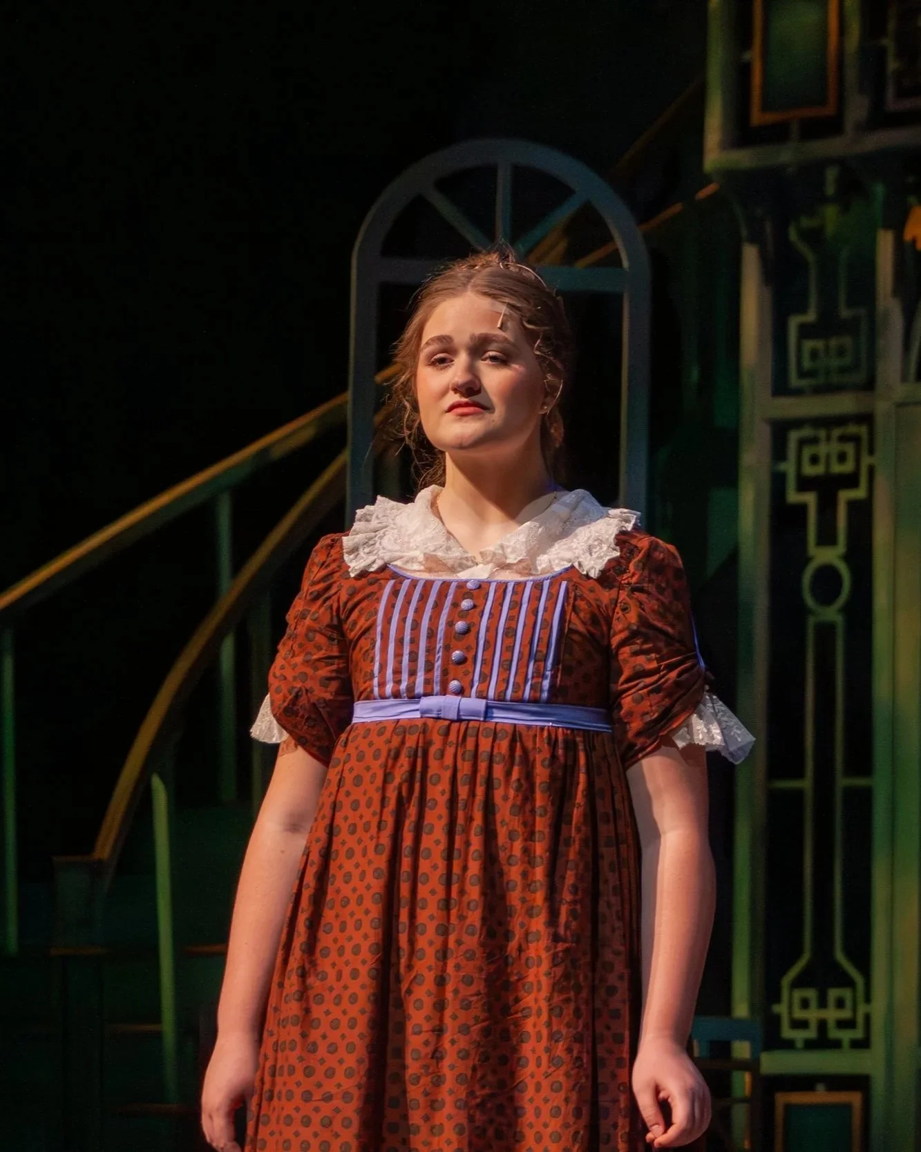 A woman in a vintage-style dress with a lace collar, standing on a stage with dark background and colorful set pieces.