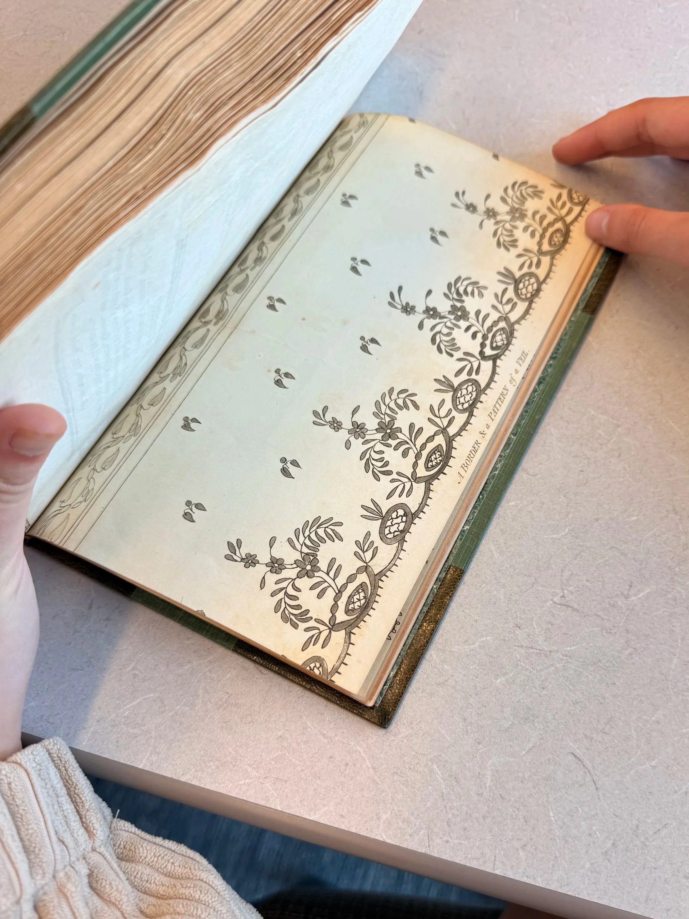 An open vintage book displaying black and white decorative border patterns on the pages, with a person's hand holding the right page down.