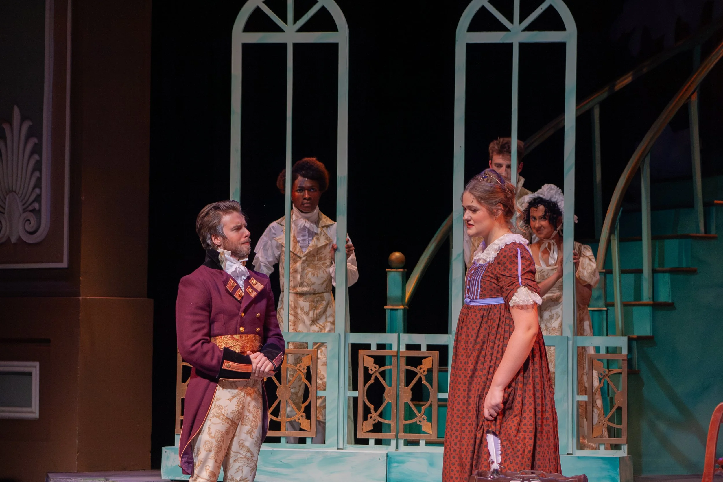 Stage scene from a theatrical play featuring five actors in period costumes engaged in a conversation, with a decorative staircase and ornate set pieces in the background.