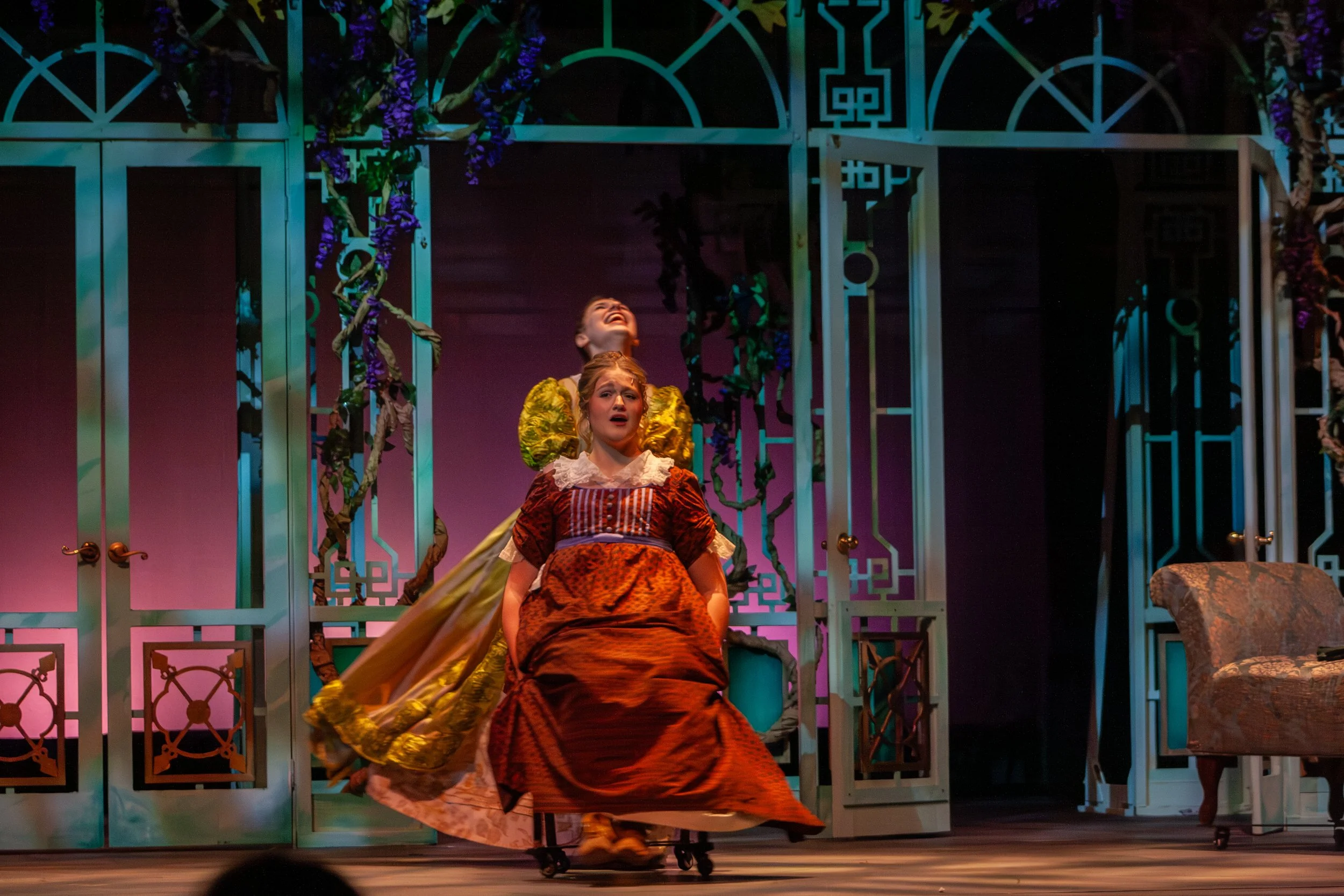 Two women on stage dressed in period costumes, one sitting and the other standing behind her. The stage background features decorative, colorful Victorian-style architecture with purple flowers and vines.