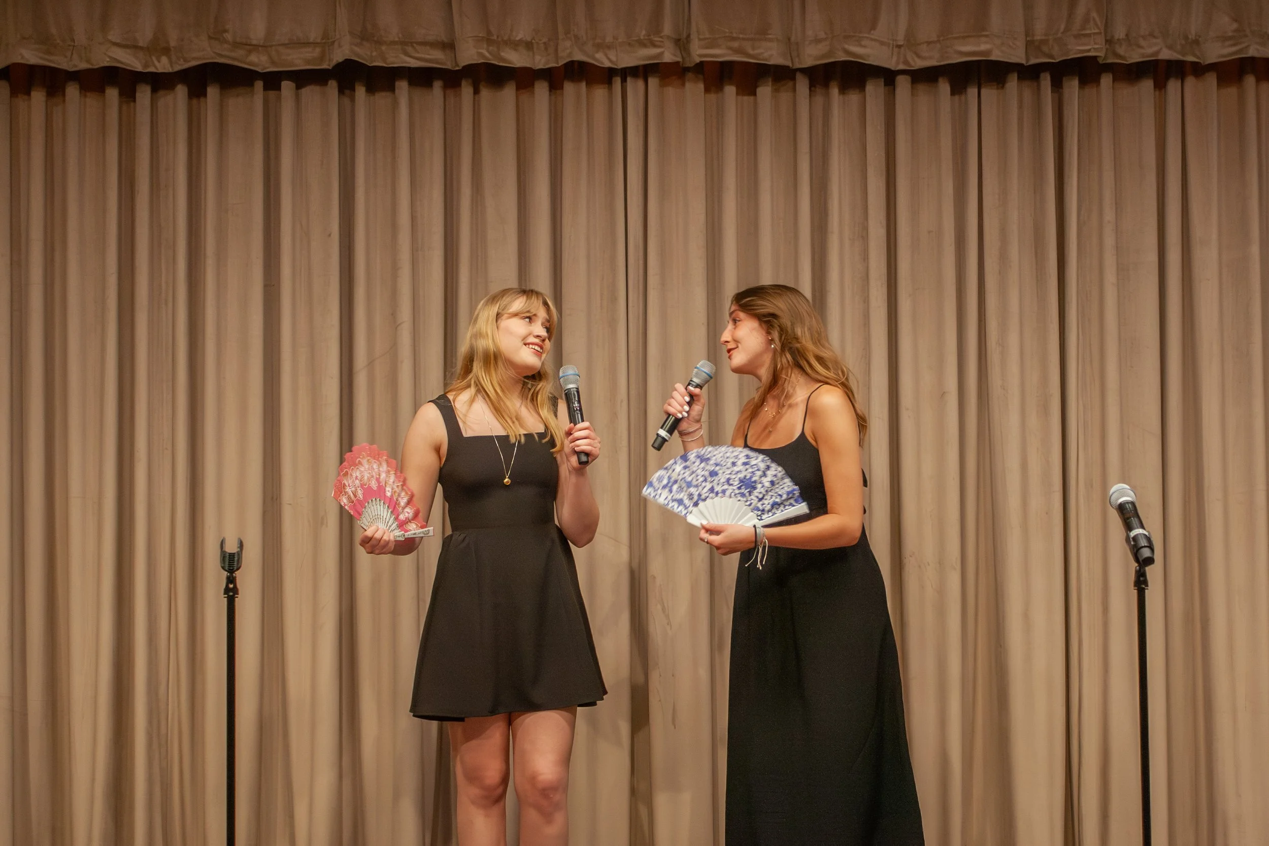 Two women are standing on a stage with a beige curtain in the background, engaging in a conversation with microphones in hand. They are dressed in black dresses and holding decorative fans, smiling at each other.