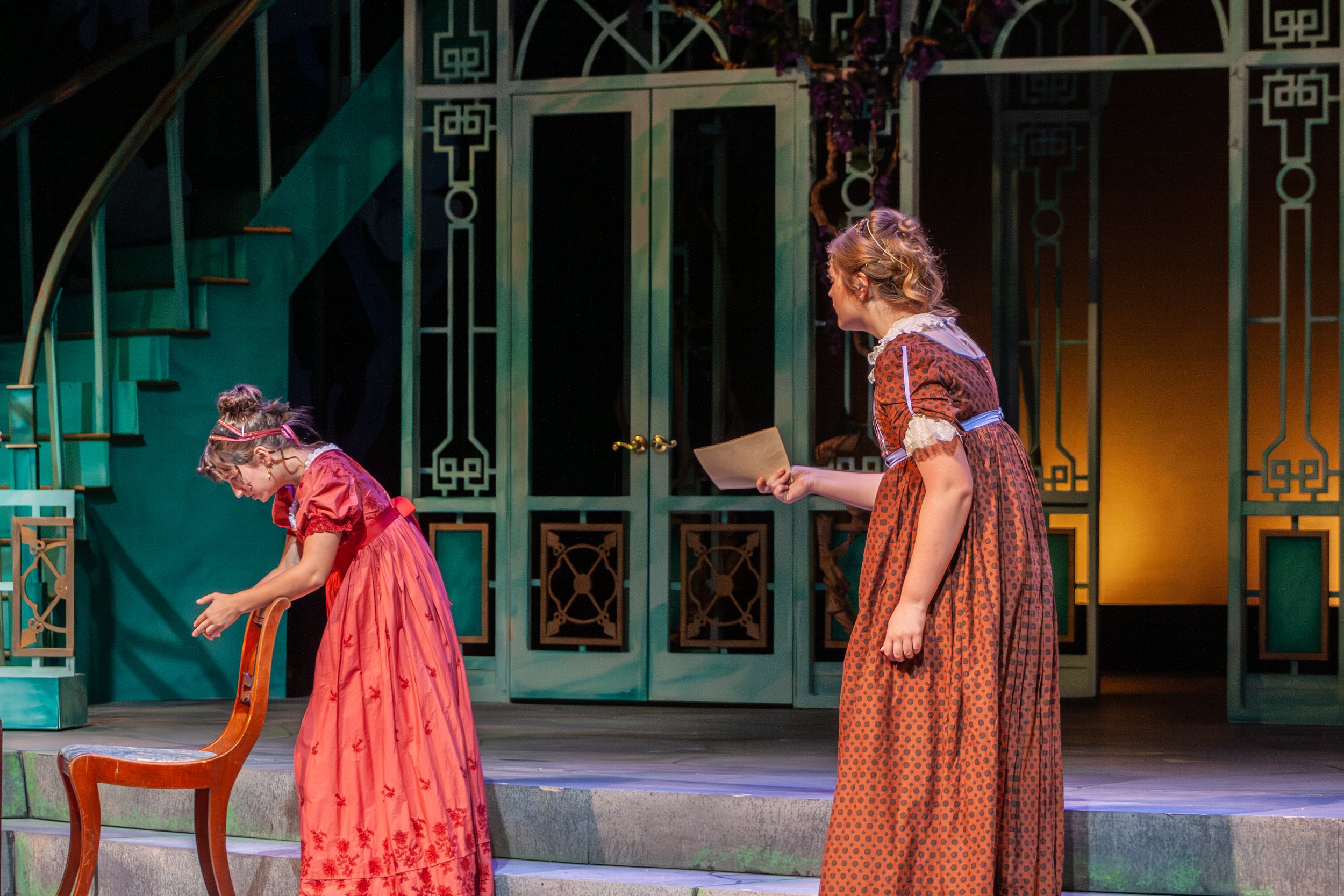 Two young women in period costumes on stage, one bowing and the other holding a script or letter, in front of a decorative set with staircase and green patterned panels.