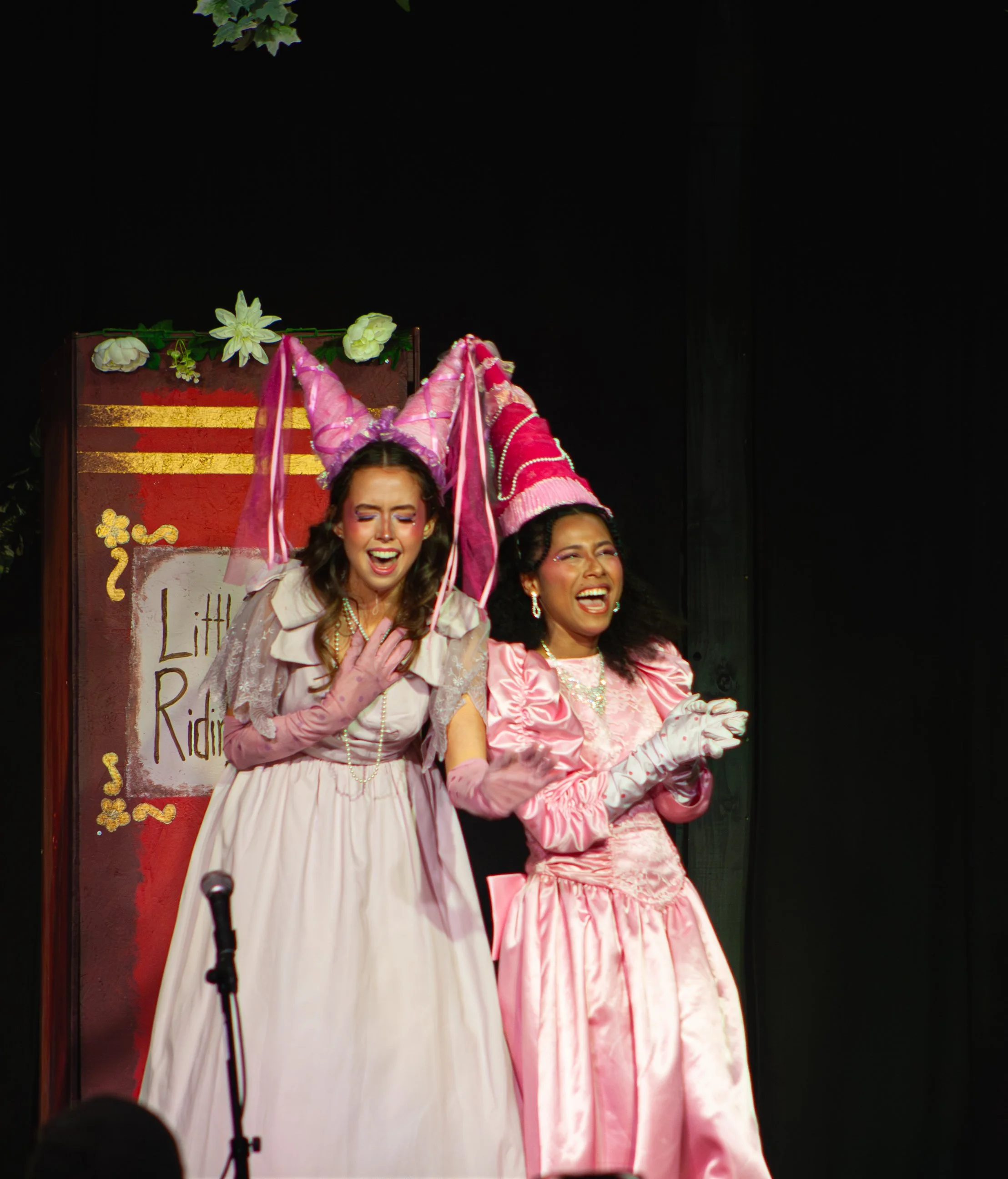Two women dressed in pink and white dresses with colorful hats and gloves, smiling on a stage with a red and gold background decorated with flowers and a sign.