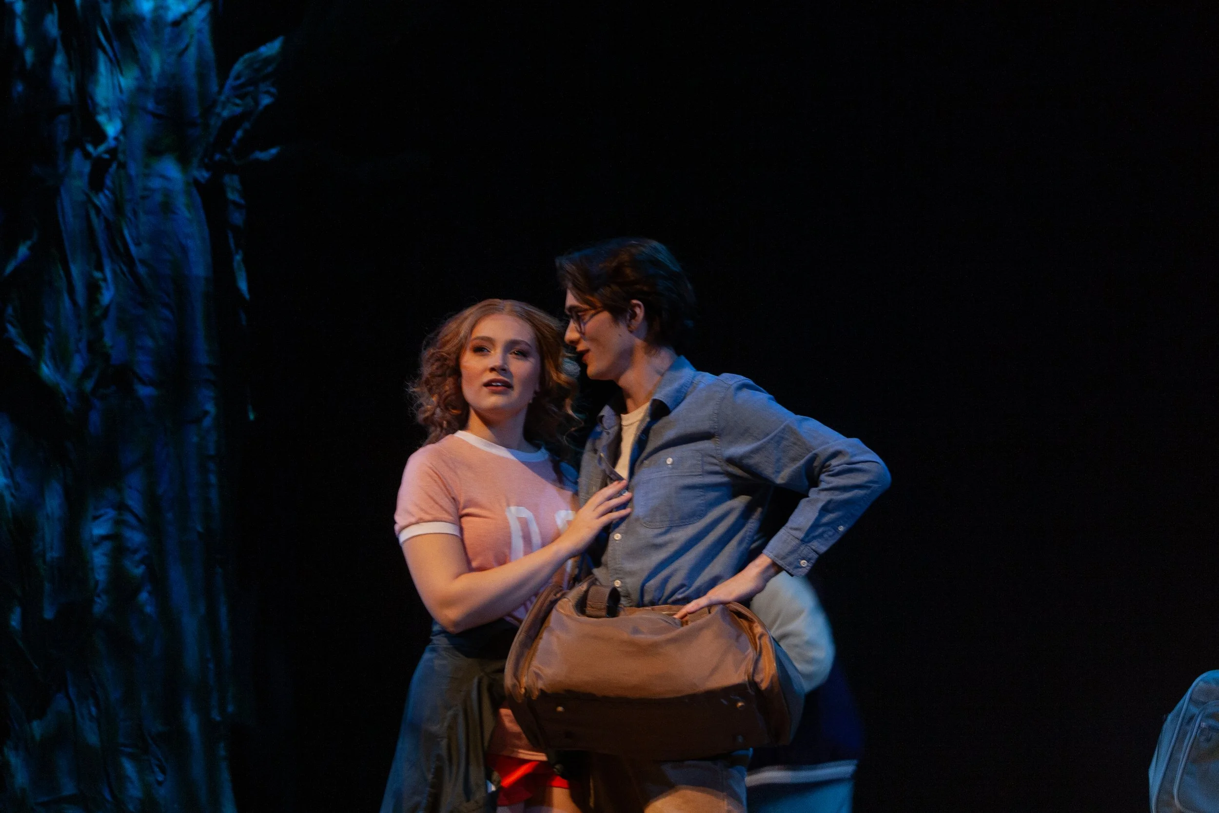 A scene from a theatrical performance featuring a woman with curly red hair and a man with dark hair and glasses, both holding a brown bag. The woman is wearing a pink and white shirt, and the man is dressed in a denim shirt. They are standing close 