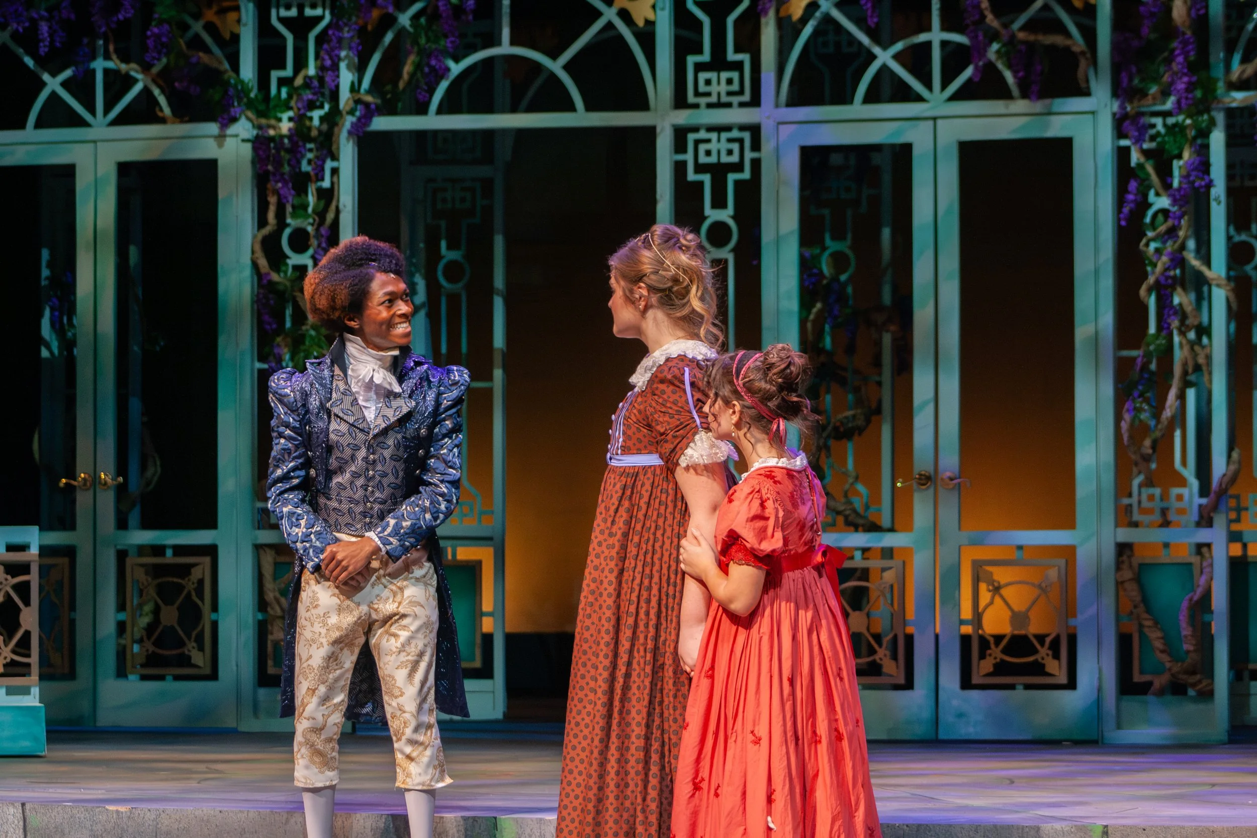 Four women in historic costumes standing on a stage with a decorative teal lattice backdrop. They are engaged in conversation, smiling, and facing each other. The scene appears to be from a theatrical performance set in a period setting.