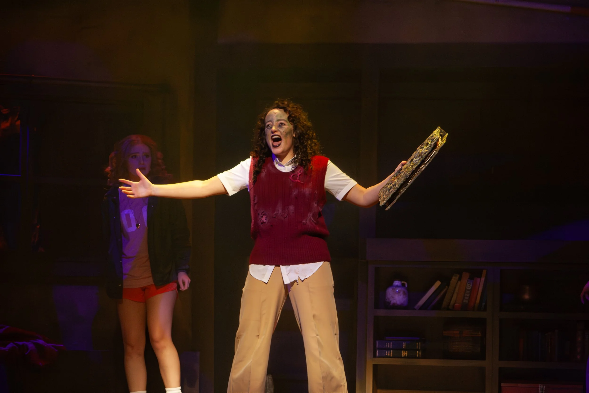 The image shows three young women standing on a stage, with one woman in the foreground holding a painting or picture. The woman in the front has curly hair and is wearing a red vest over a white shirt with frayed beige pants. She appears to be singi