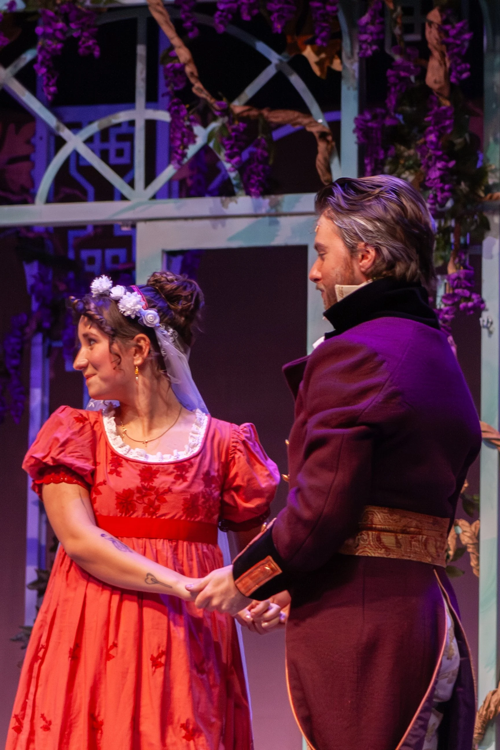 A man and woman standing close, holding hands and looking at each other during a theatrical performance, with purple flowers and decorative lattice in background.
