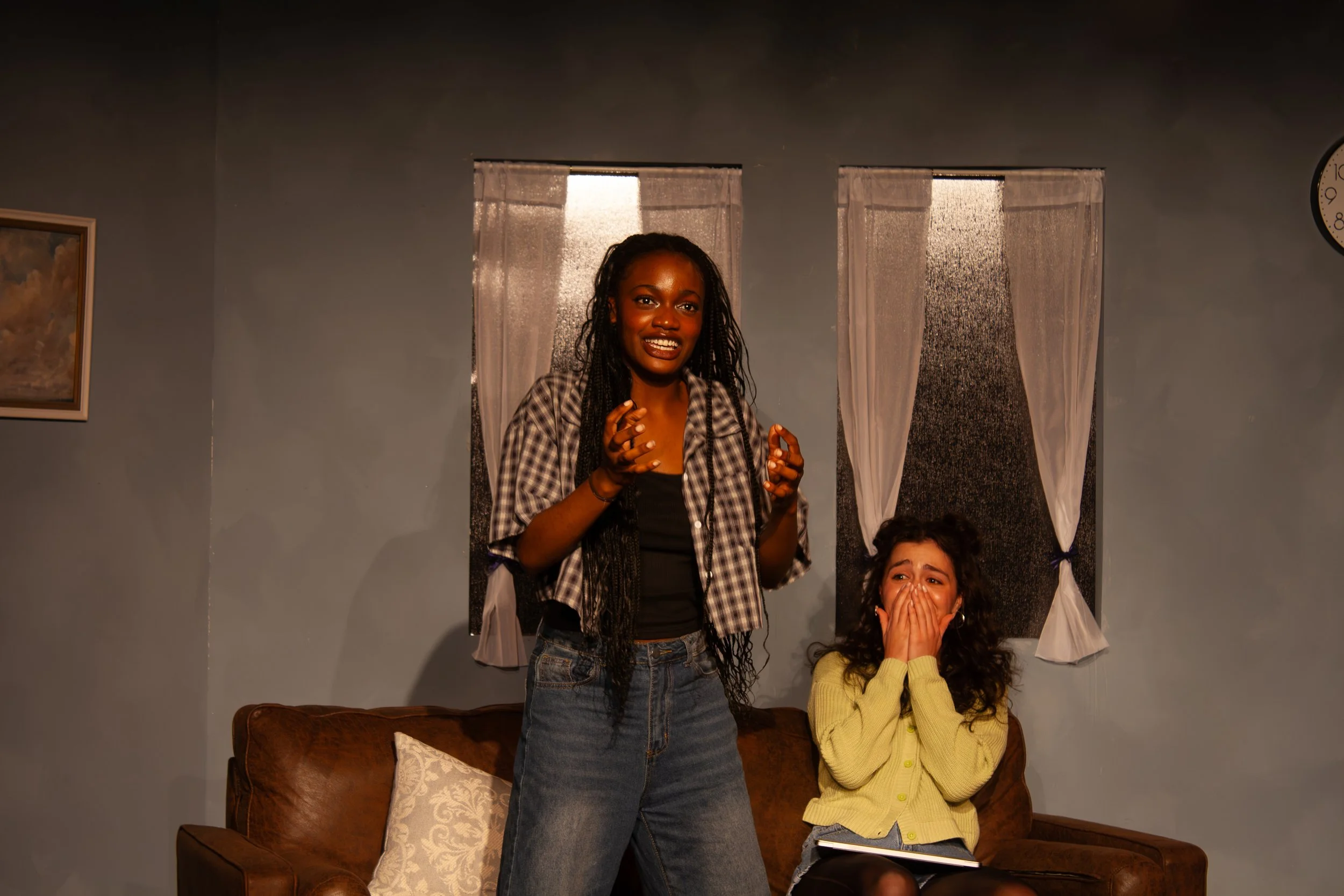 Two women in a living room, one standing and one sitting on a brown leather couch, with a wall clock and framed picture in the background. The woman standing has a surprised or excited expression, and the woman sitting covers her mouth with her hands