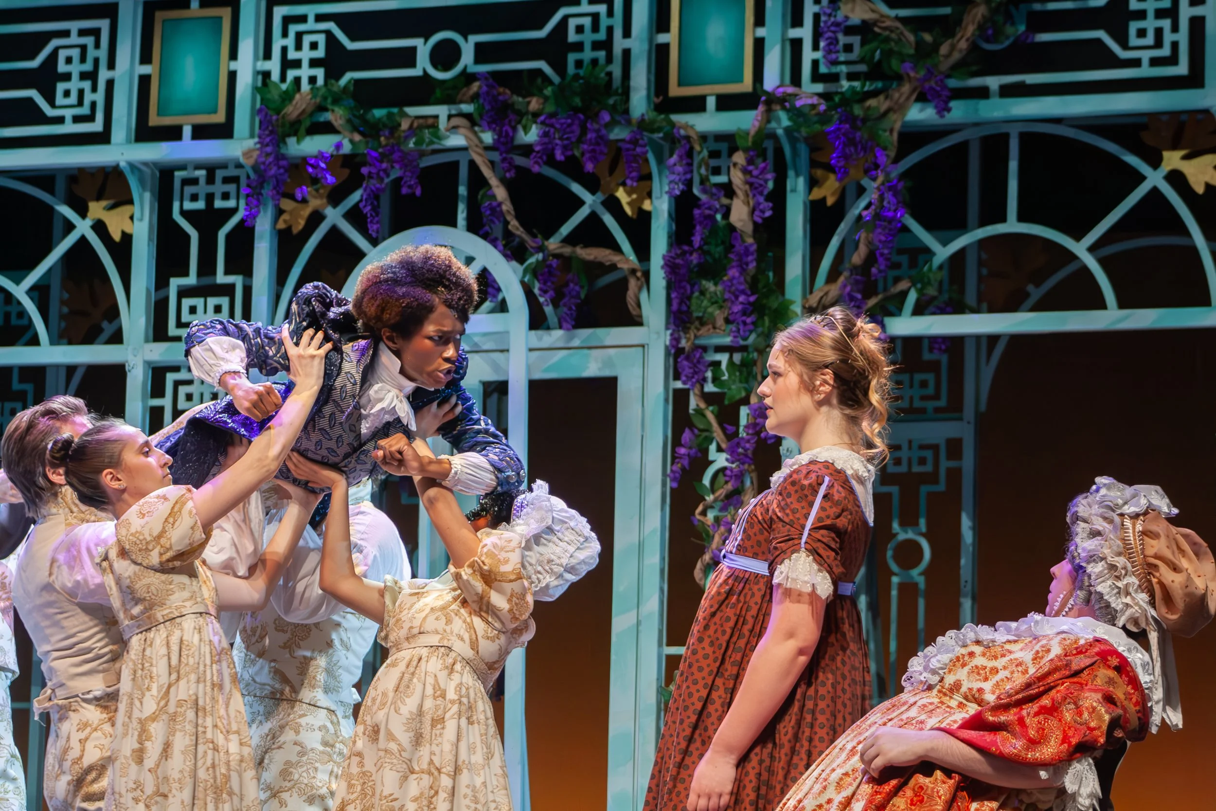 Scene from a theatrical play featuring actors in historical costumes, with a colorful set design including purple flowers and intricate fencing.