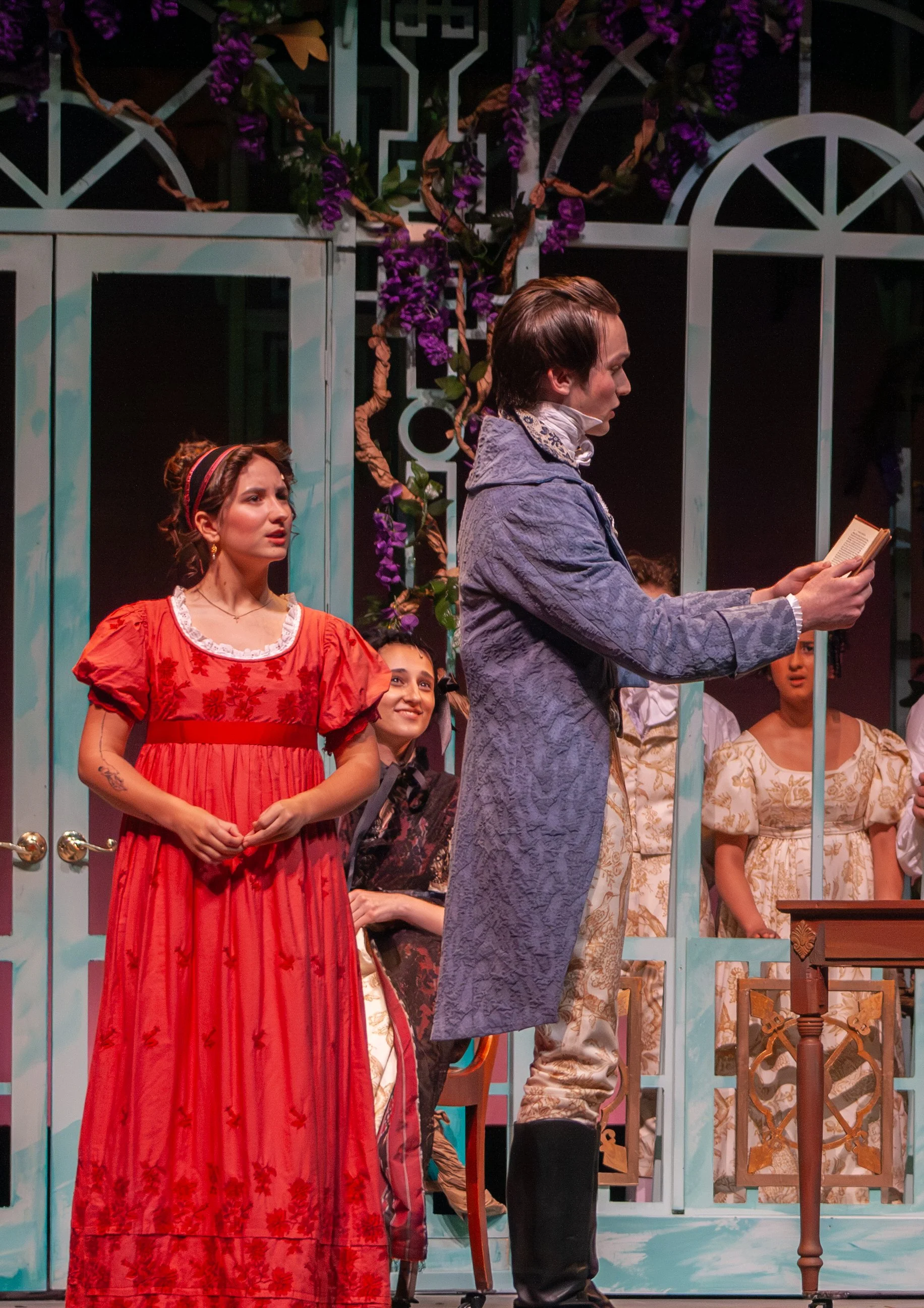 The scene appears to be part of a theatrical performance with actors dressed in period costumes, standing in front of a decorative backdrop with purple flowers and garden-like fencing. A young woman in a red dress looks on, while a young man in a pur