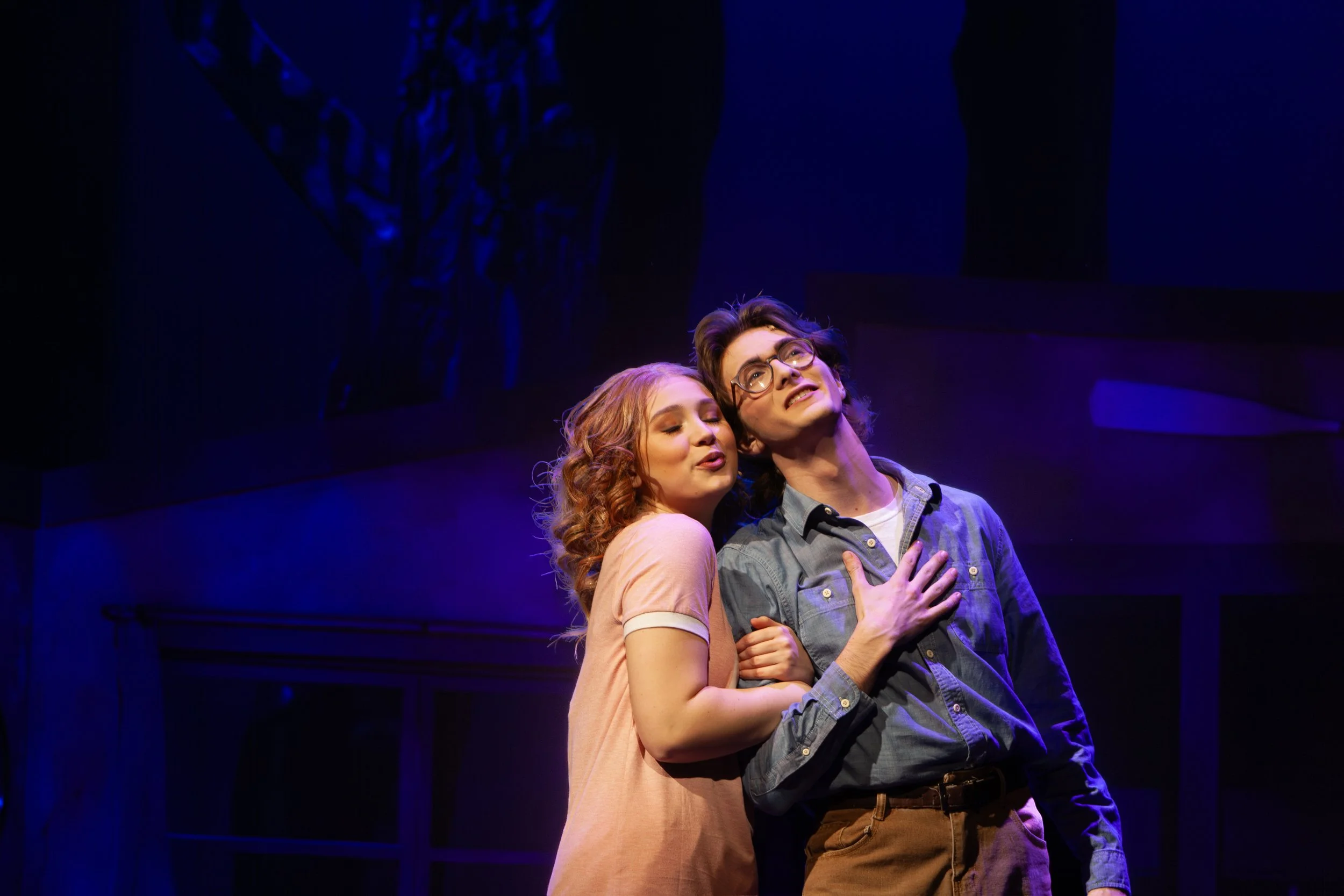 A young woman with curly red hair and a young man with glasses and dark hair are on stage, close together. The woman is leaning on the man's chest with her eyes closed, while the man has one hand on his chest and is looking upward with an emotional expression. They are bathed in purple stage lighting.