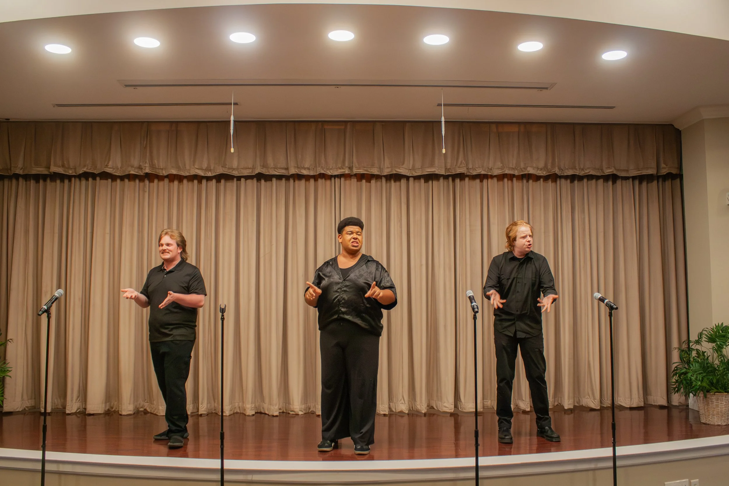 Three people are performing on a stage with a beige curtain backdrop. They are standing in front of microphones, wearing black attire, and appear to be engaged in a musical or theatrical presentation.
