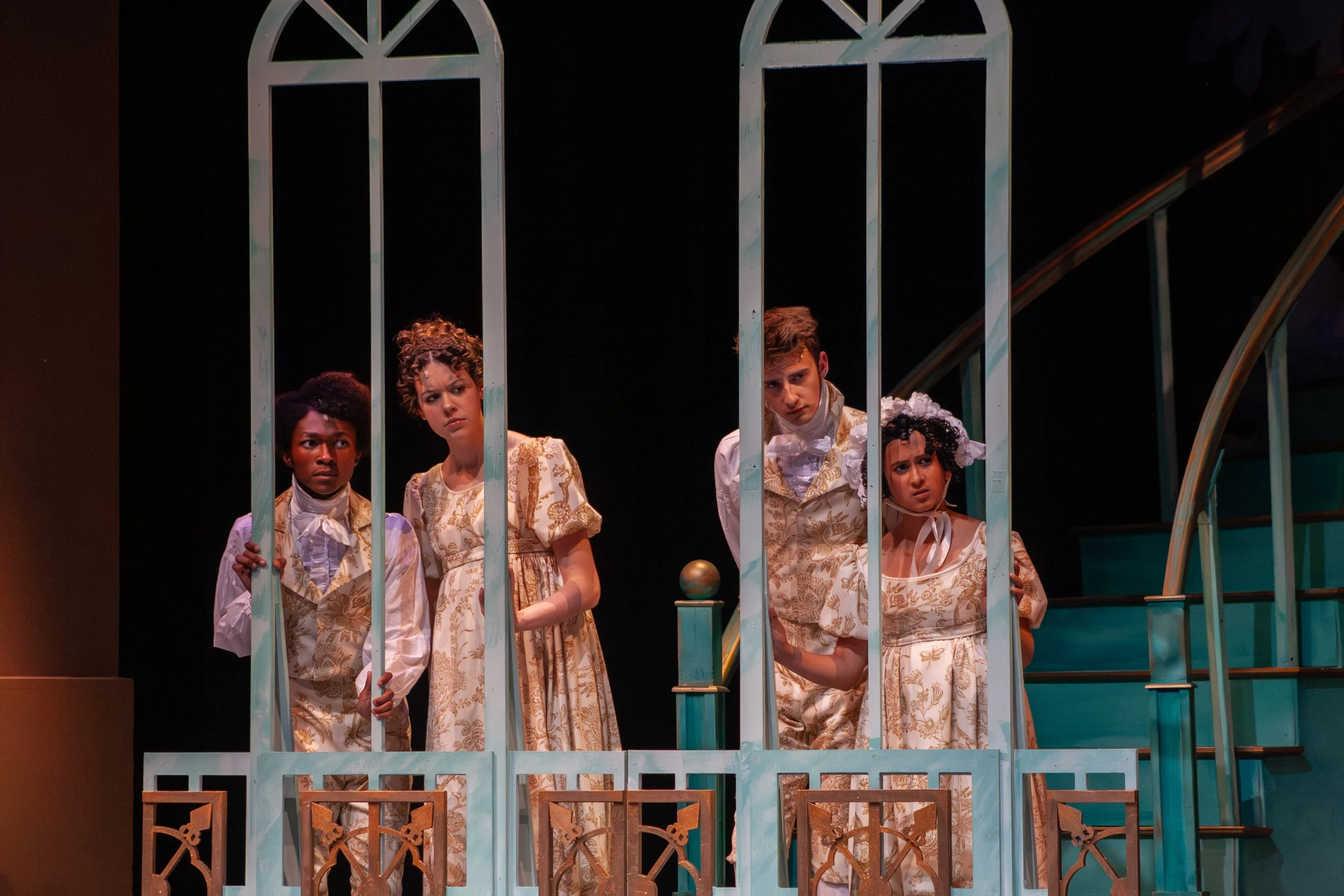 Four children wearing vintage-style dresses and coats look through the bars of a decorative teal-colored fence or gate on a stage, possibly during a theatrical performance.