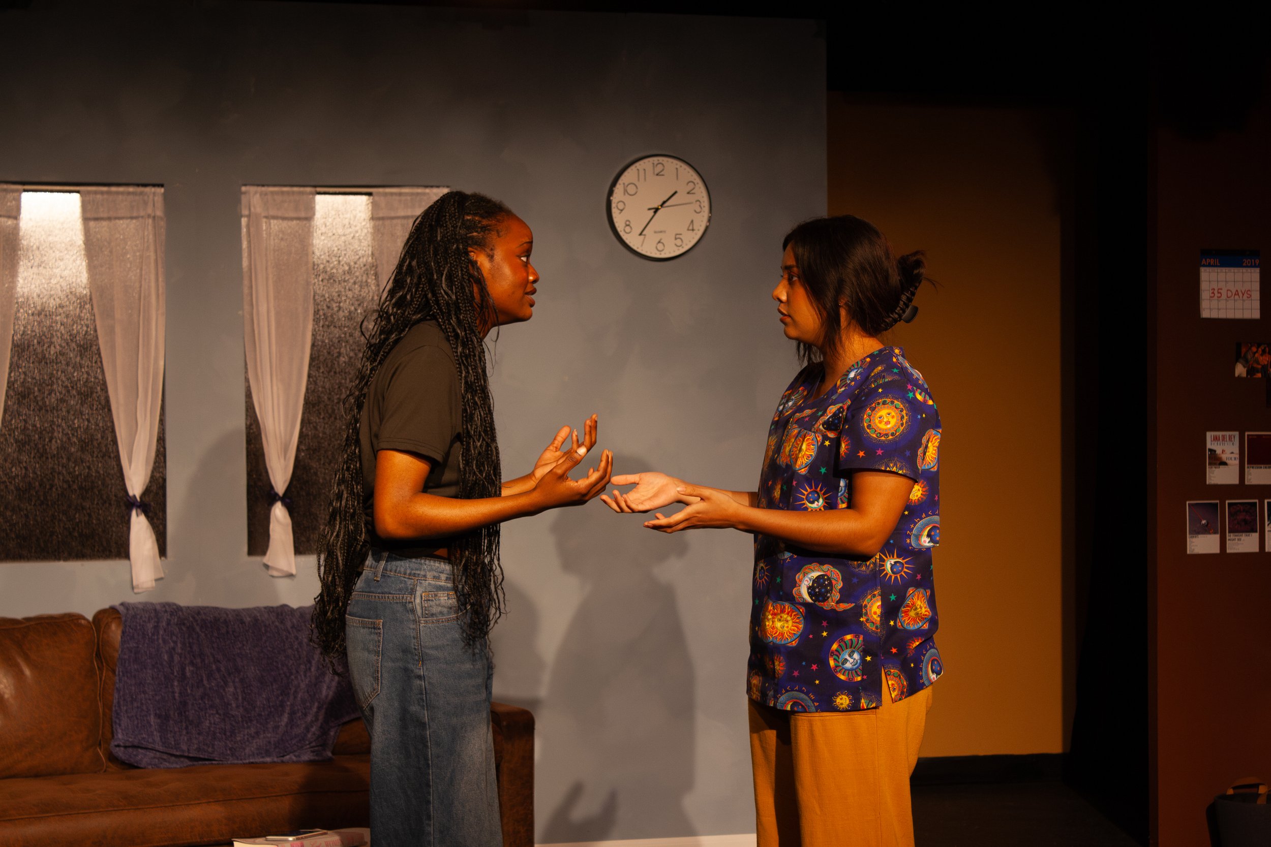 Two women stand facing each other holding hands and talking in a living room with a gray wall, a clock, two windows, and a bookshelf.