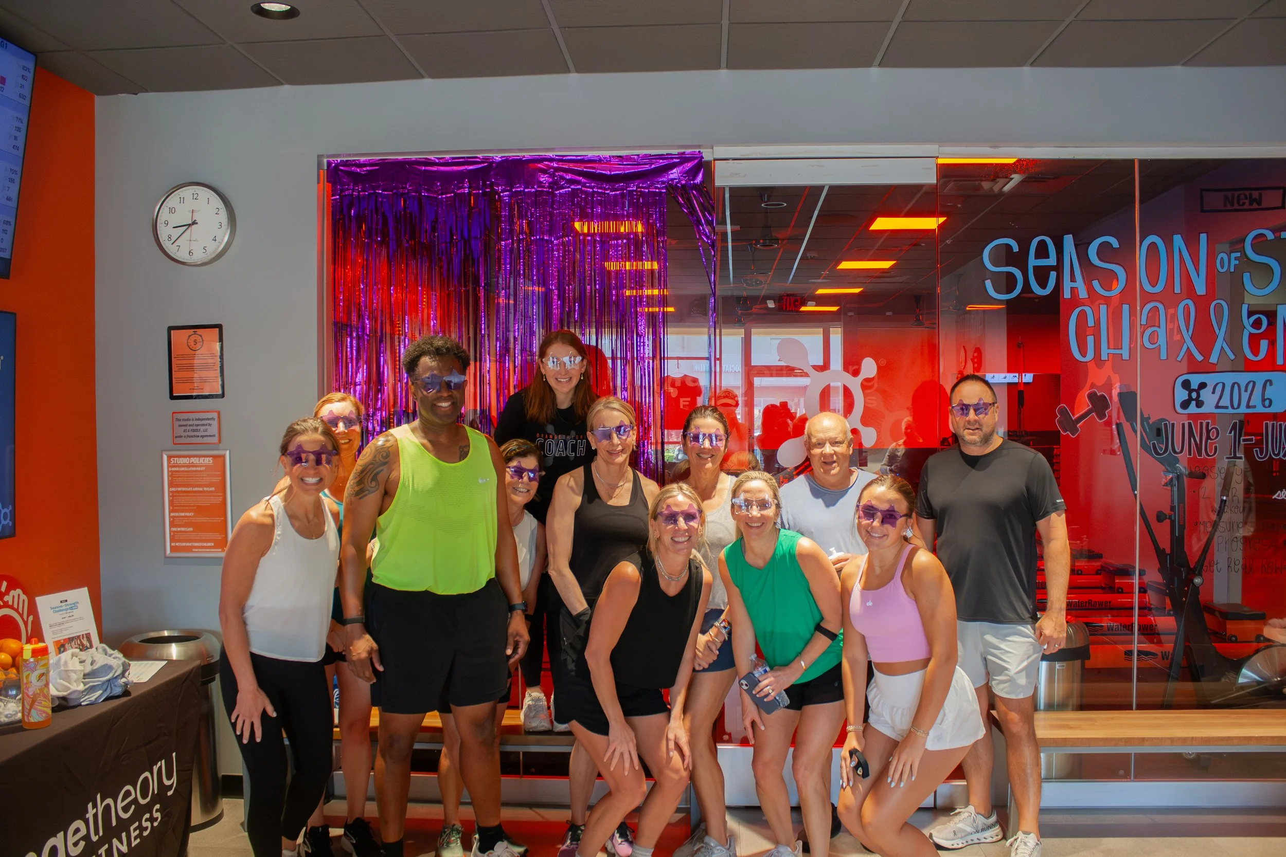 A group of smiling people wearing sunglasses and athletic clothing gathered inside a fitness center next to a brightly lit sign that reads 'Season of Challenge 2026'
