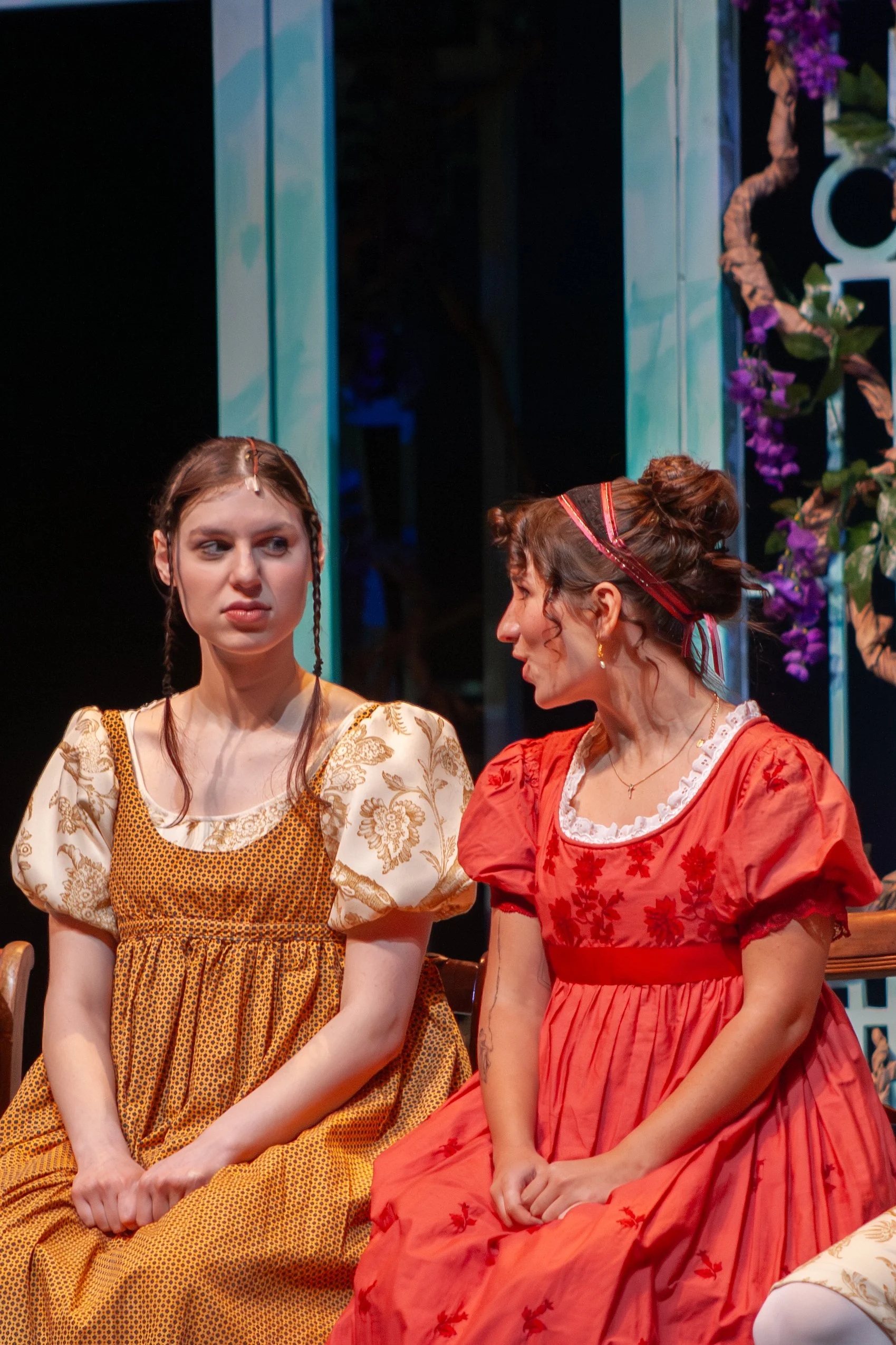 Two women dressed in historical costumes sit and engage in conversation on a stage, with a backdrop of floral decorations and vertical teal-colored panels.