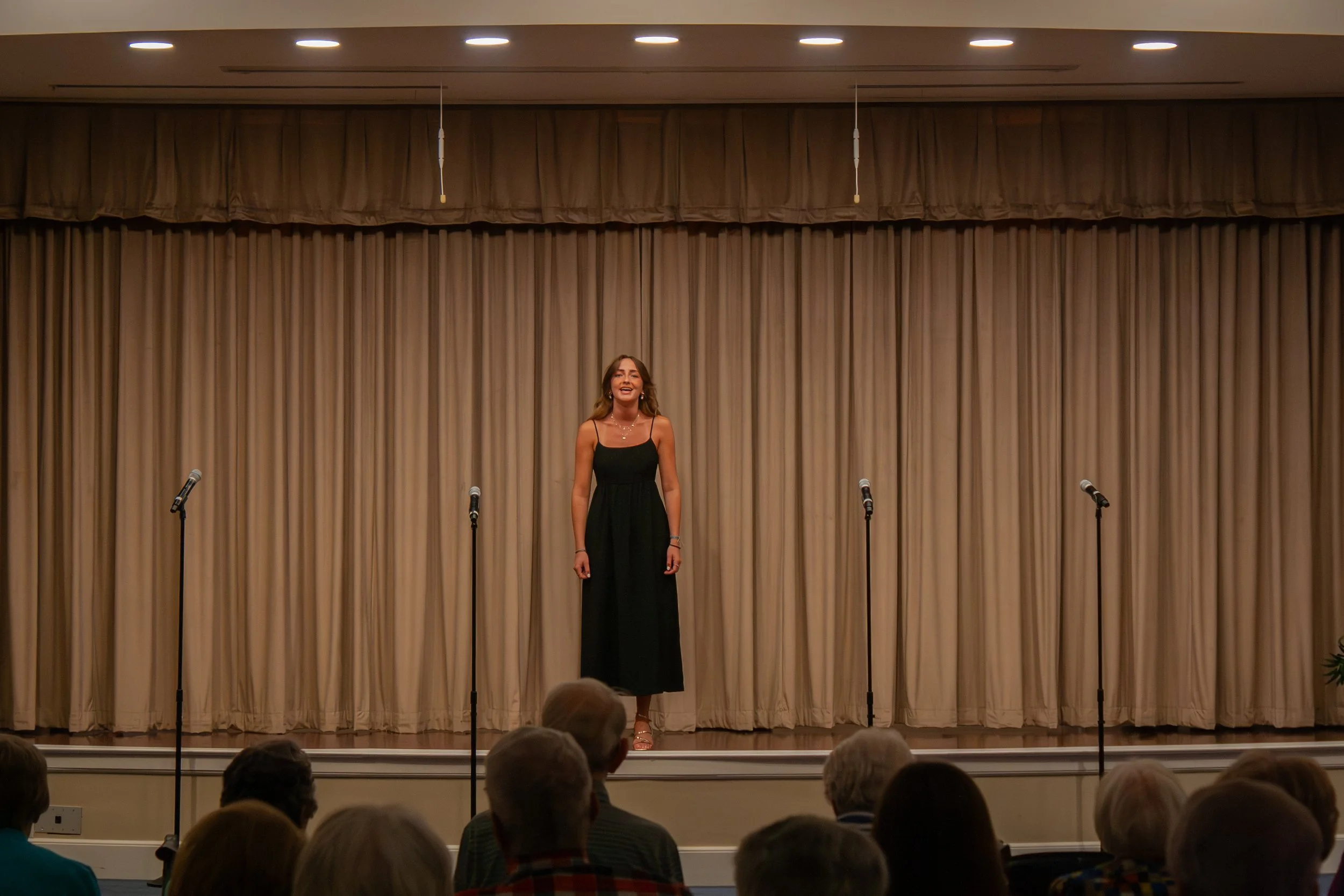 A woman in a black dress standing on a stage in front of a beige curtain, speaking or singing into a microphone. Audience members are visible in the foreground.