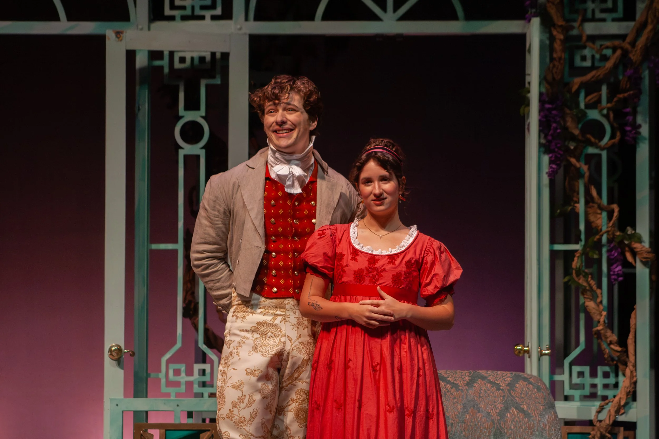 Two actors on stage in period costumes, standing in front of a decorative garden gate with purple flowers, during a theatrical performance.