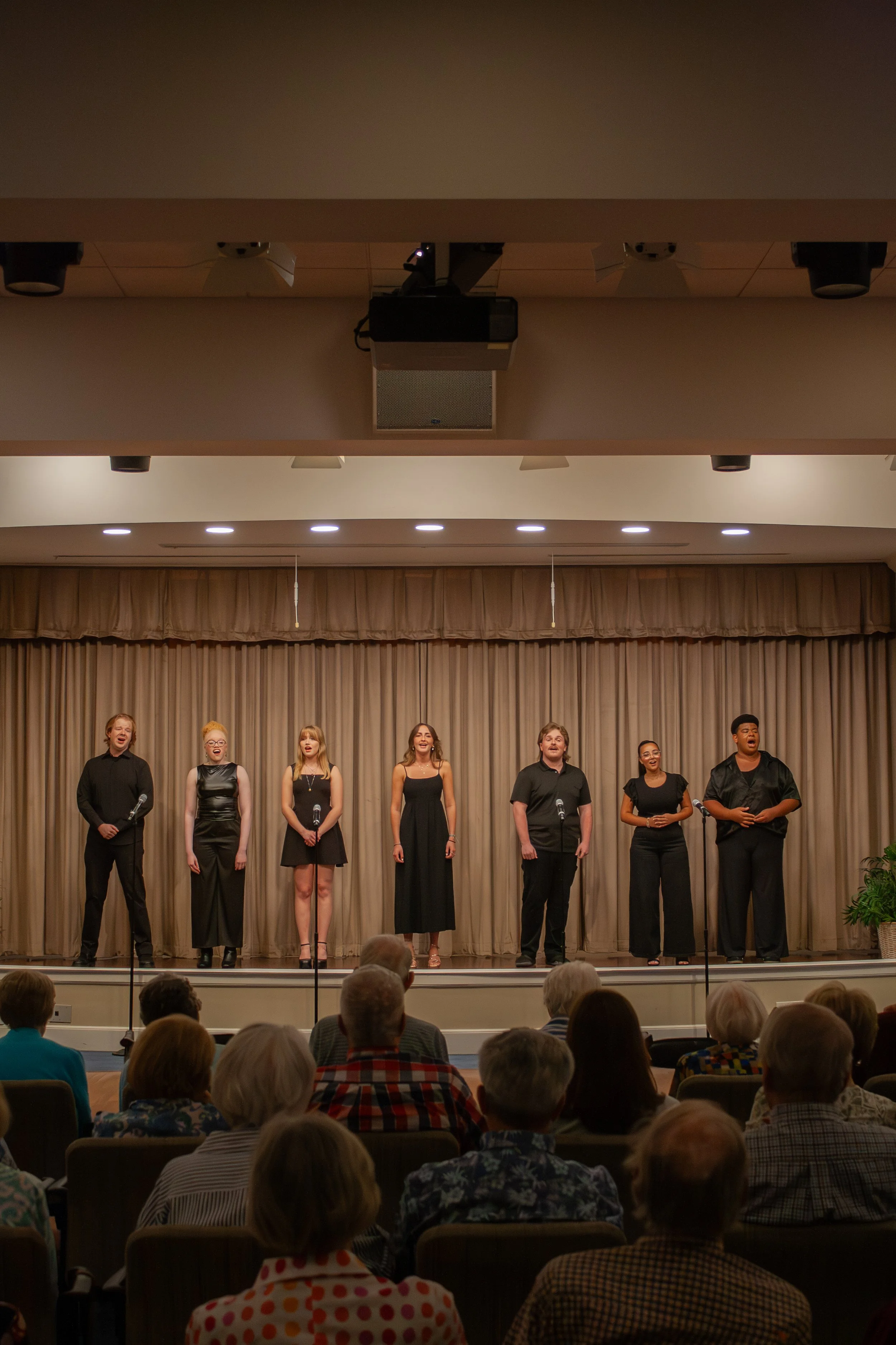 A choir of diverse individuals singing on stage in front of an audience with beige curtains and warm lighting.