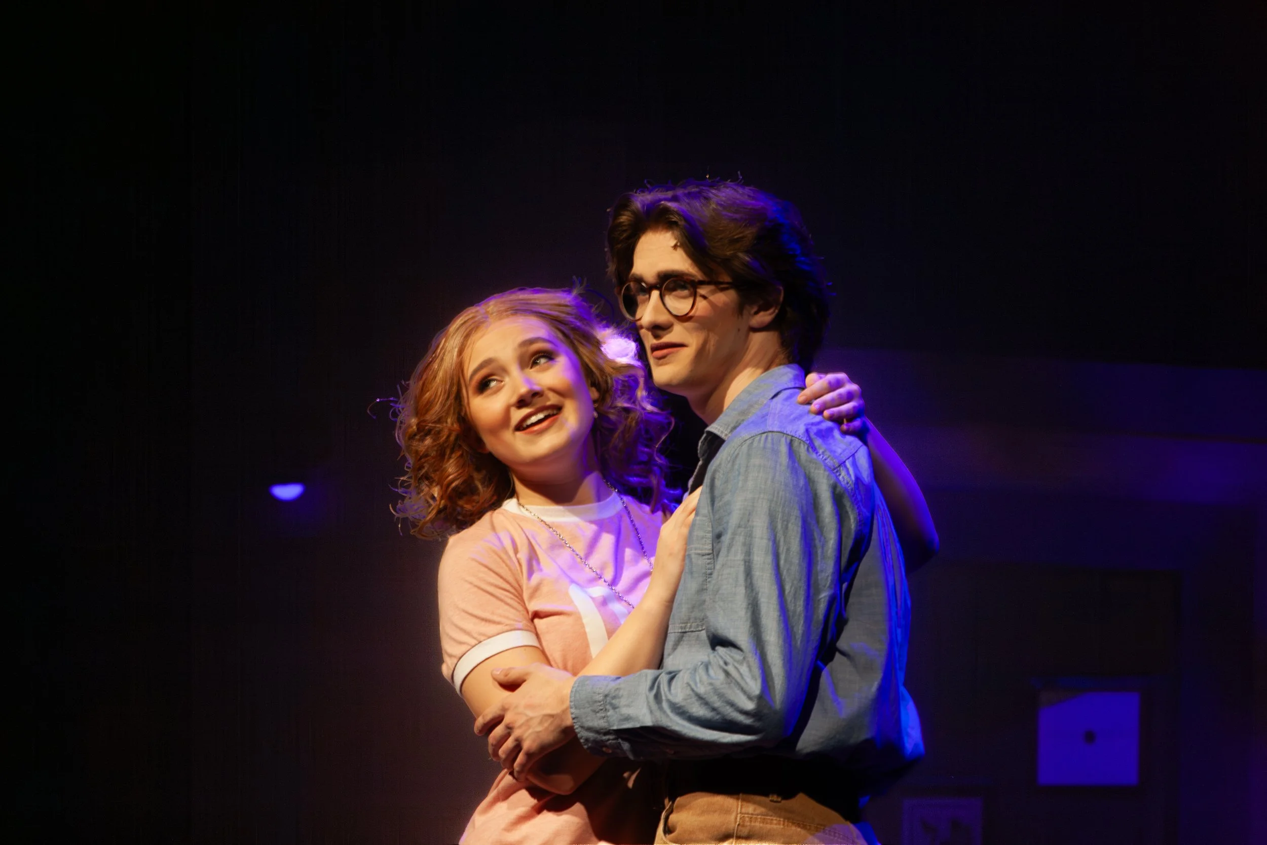 A young woman and a young man are dancing together in a dimly lit setting, with the woman smiling and looking up at the man. The woman has red hair, and the man has dark hair, glasses, and is wearing a light blue shirt. They appear to be enjoying a m