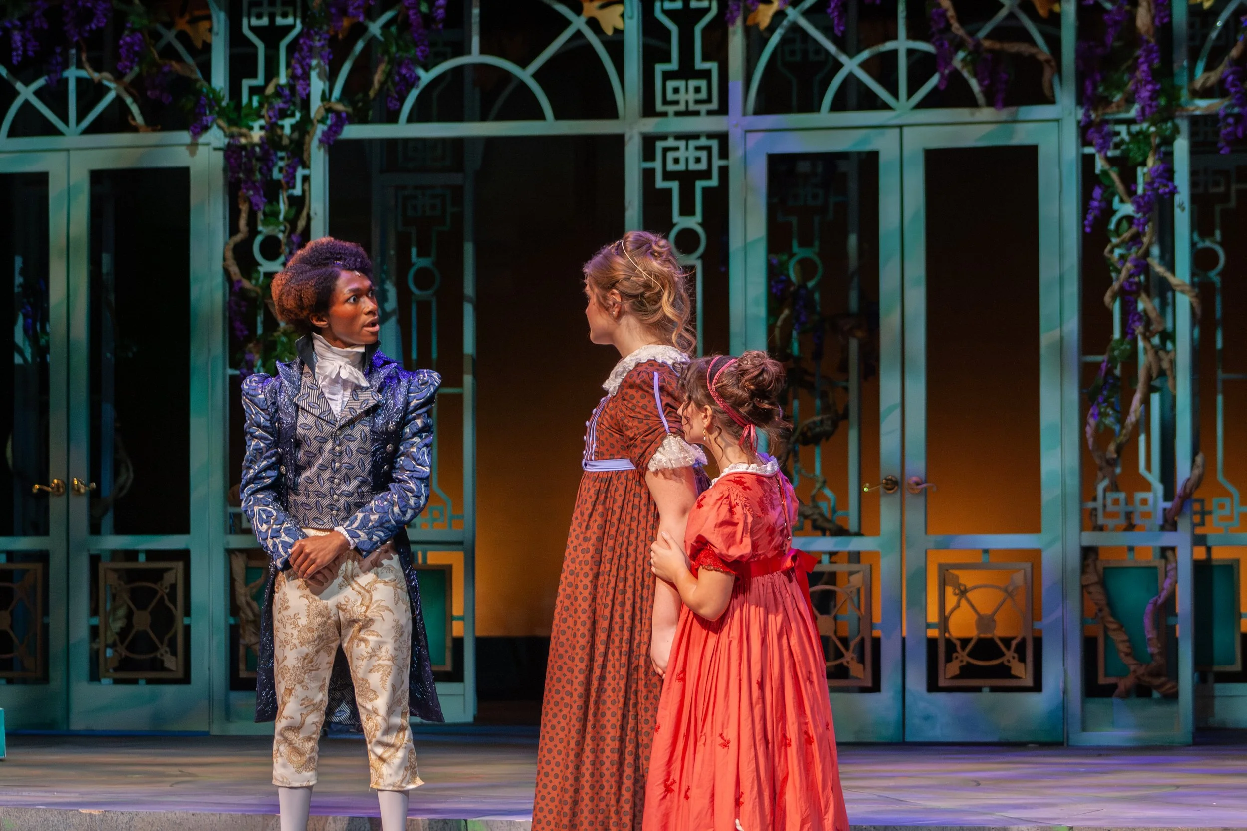 Four women in period costumes standing and facing each other on a theater stage with a decorative teal gate and purple flowers in the background.