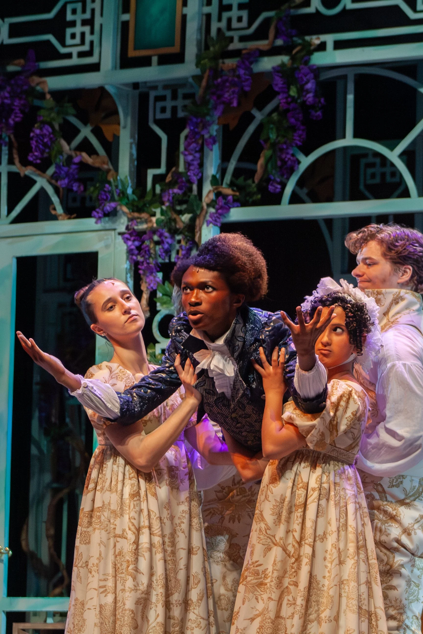 The image depicts a theatrical scene with five performers dressed in period costumes, with three young women and two young men, on stage with a decorative backdrop featuring purple flowers and ornate design elements.