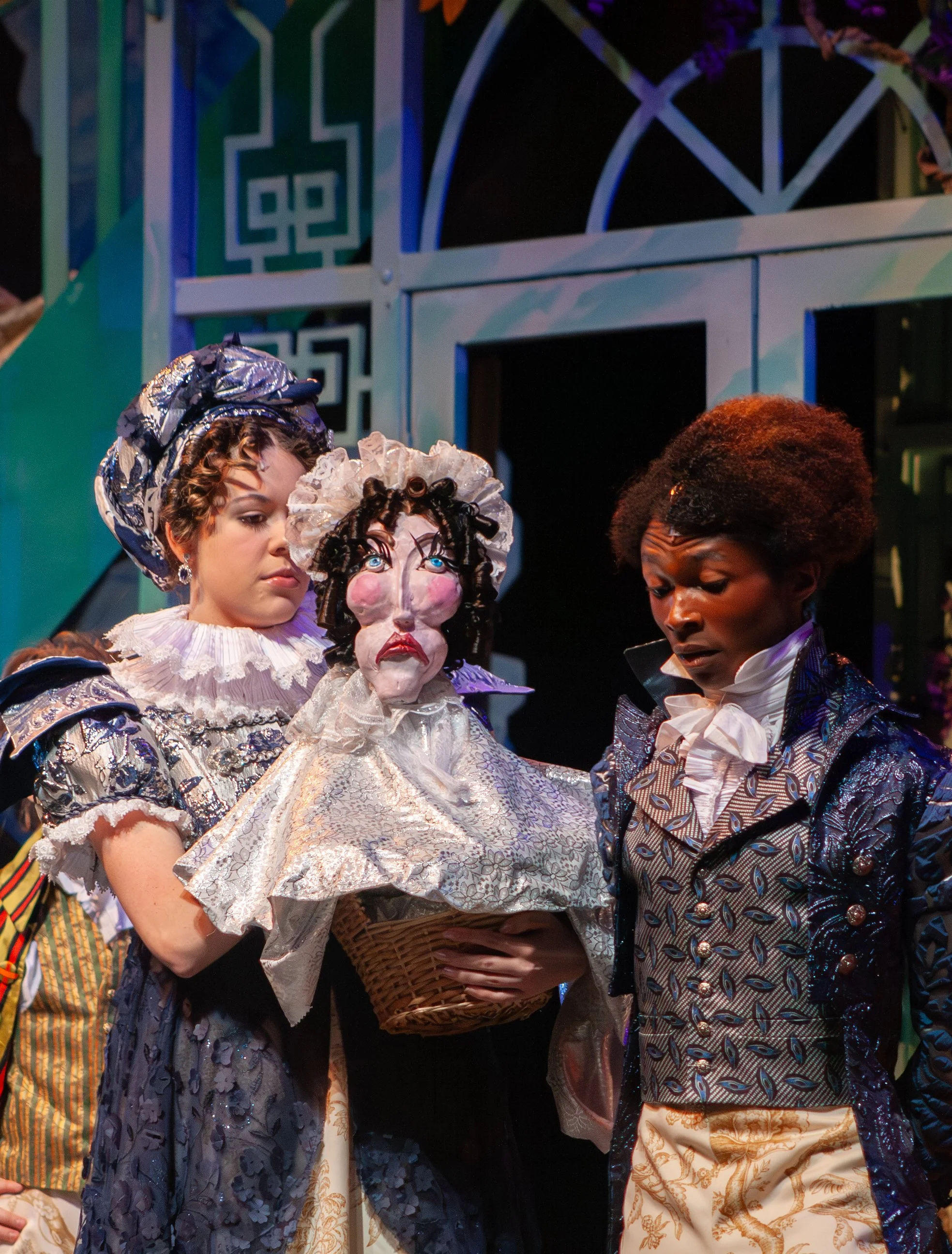 Three people in elaborate historical costumes, with a background of decorative architectural elements that include geometric patterns. One person is holding a basket.